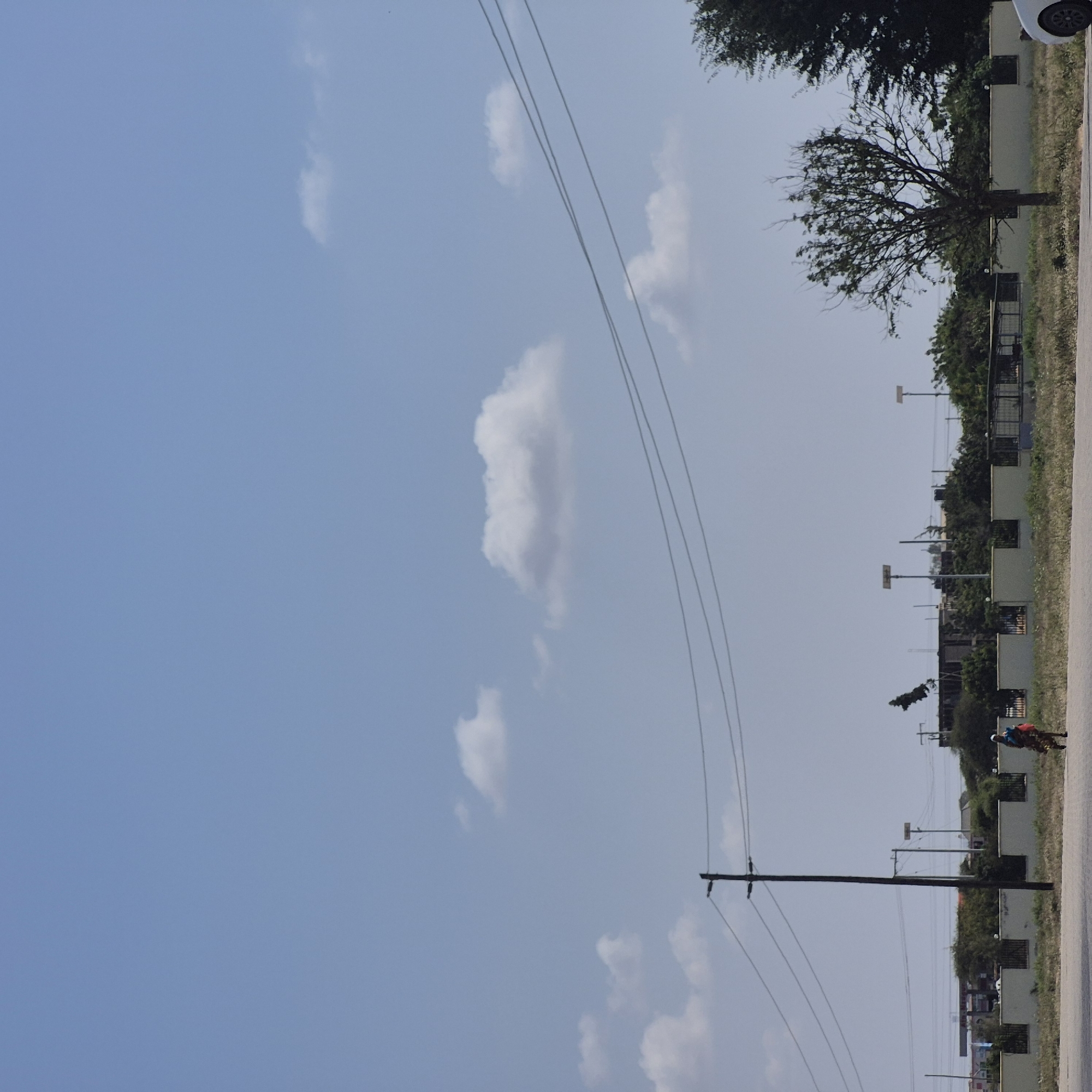 The image depicts a daytime scene in Dodoma, Tanzania. The sky is a clear, light blue with scattered white clouds. In the foreground, a dirt road or pathway runs horizontally across the bottom of the frame. To the left, a dark brown utility pole stands tall, supporting multiple power lines that stretch across the image.
Further back, a person, appearing to be a woman based on her attire, walks from left to right, carrying a bundle or bag. She is wearing a colorful patterned dress and a head covering. Behind her, a low, cream-colored wall with a dark metal fence runs parallel to the road. Beyond the wall, various structures are visible, including what appears to be a gas station with visible signage and pumps, several buildings with roofs, and an assortment of trees and vegetation. Two tall, slender poles with solar panels are also noticeable in the background. To the far right, the branches of a large, bushy tree are visible, and a white car is partially in the frame. The overall environment suggests an urban or semi-urban setting with infrastructure and residential or commercial development.