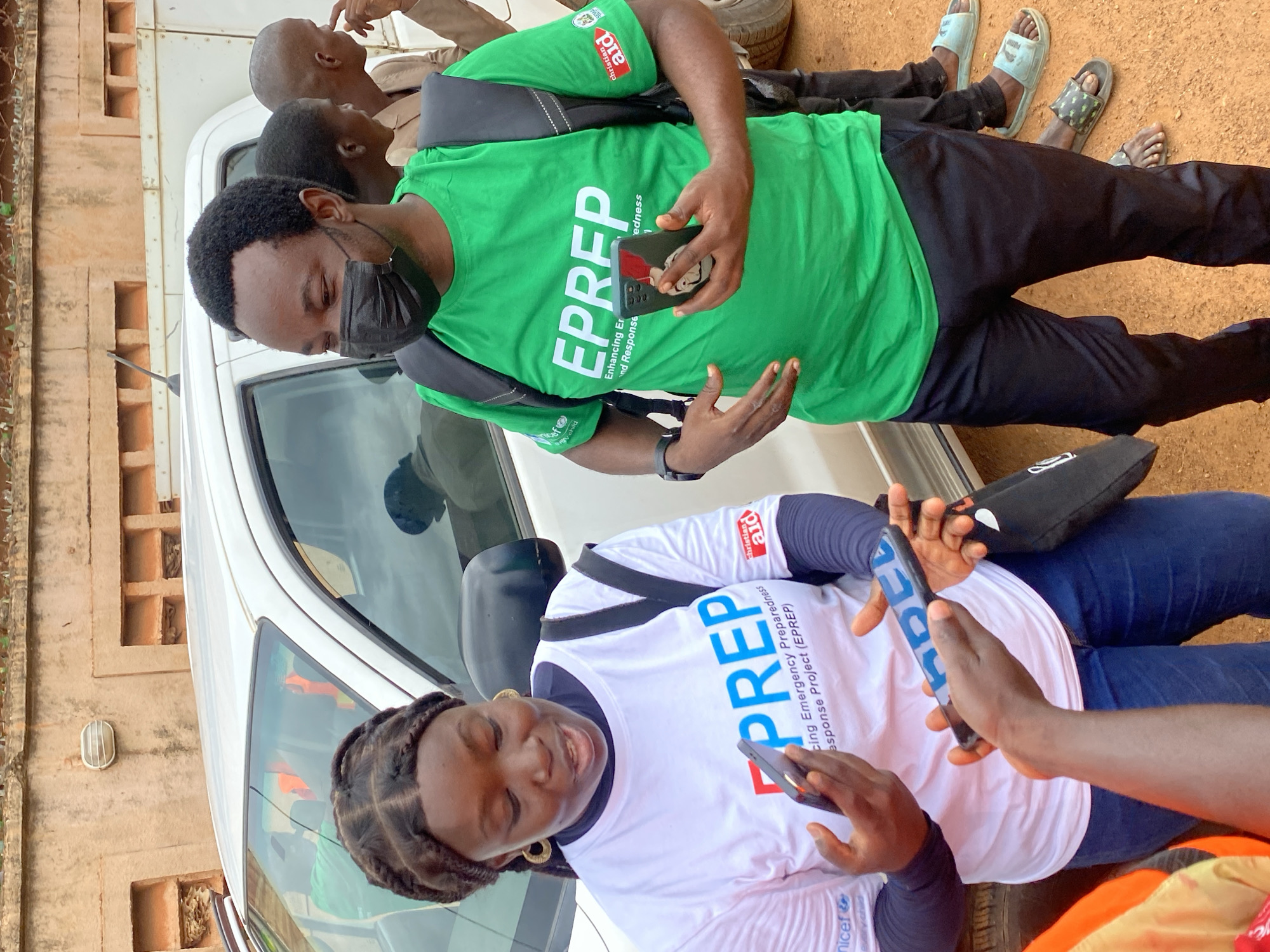 In Kawo, Nigeria, a woman with braided hair and gold hoop earrings, wearing a white long-sleeved shirt with blue and red text that reads "EPREP Enhancing Emergency Preparedness and Response Project (EPREP)" and a UNICEF logo, is holding a dark-colored smartphone and talking to someone off-camera. A man wearing a black face mask and a green t-shirt with white text that reads "EPREP Enhancing Emergency Preparedness and Response" and Christian Aid and other logos, is standing to her right. He holds another dark-colored smartphone in his right hand and has a black backpack on. Behind them, a white vehicle is parked. The ground is covered in reddish-brown dirt, and a textured brick wall with small alcoves and an outdoor light fixture is visible in the background.