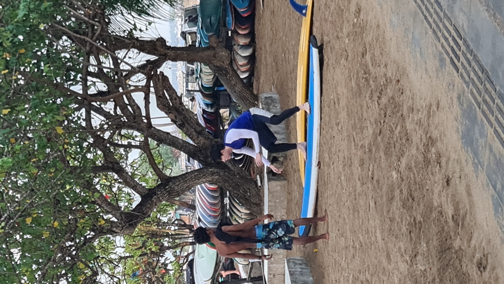 The image depicts a sandy beach environment in Denpasar, Indonesia, with numerous surfboards stacked in the background and a rack of surfboards visible to the right. In the foreground, two individuals are present. One person, wearing black leggings and a blue and white long-sleeved shirt, is on a blue surfboard, positioned in a crouched stance as if practicing surfing. The other person, standing to the left, is shirtless and wearing patterned shorts, facing the individual on the surfboard. A tree with branches extends from the upper left quadrant of the image, providing shade. The ground is sandy, with a paved walkway visible in the lower right corner.