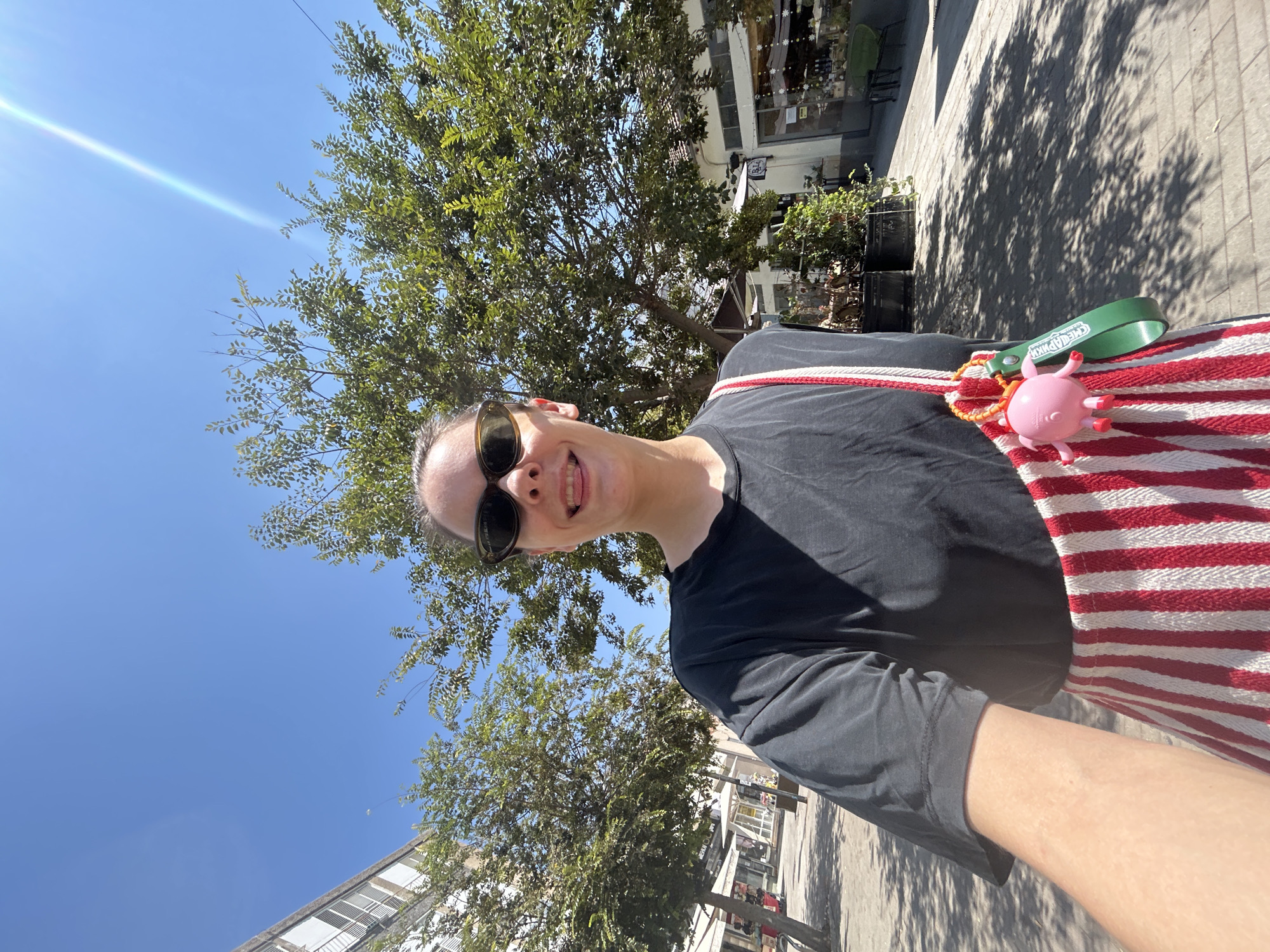 A young woman with dark hair pulled back is taking a selfie outdoors in a city. She is wearing dark sunglasses and a black t-shirt, and a red and white striped bag with a pink pig charm hangs on her shoulder. She is sticking her tongue out playfully. The background shows trees and buildings along a paved walkway, suggesting a commercial area. The image appears to be taken on a sunny day. The location is the city of Rishon LeZion, Israel, likely on Rothschild Street, a well-known pedestrian promenade lined with trees and shops.