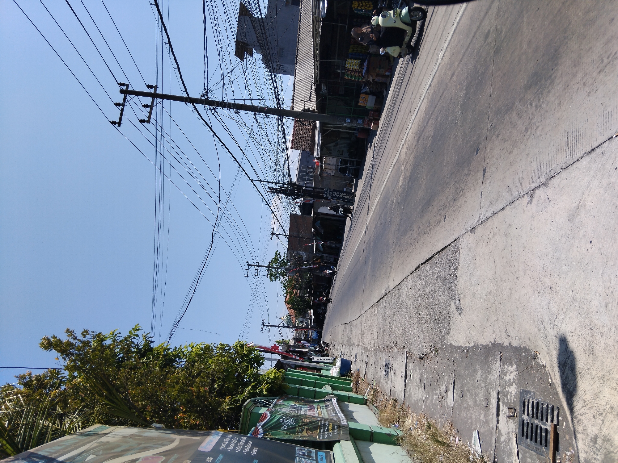 The image depicts a street scene in Kepuh Kiriman, Indonesia, under a clear blue sky. The road is paved and appears to be an urban or semi-urban environment. On the left side of the road, there is a sidewalk with some vegetation and a green fence. Several posters are attached to the fence. Power lines crisscross the sky, supported by utility poles.
Several buildings line the right side of the road, some appearing to be shops or small businesses. One building has a corrugated metal roof. A light-colored scooter with a rider is visible on the road, moving away from the viewer. Further down the road, other motorcycles and what appears to be a person on foot are present, suggesting moderate activity. The overall impression is a typical day in a populated area of Indonesia, with the presence of infrastructure like power lines and commercial establishments.