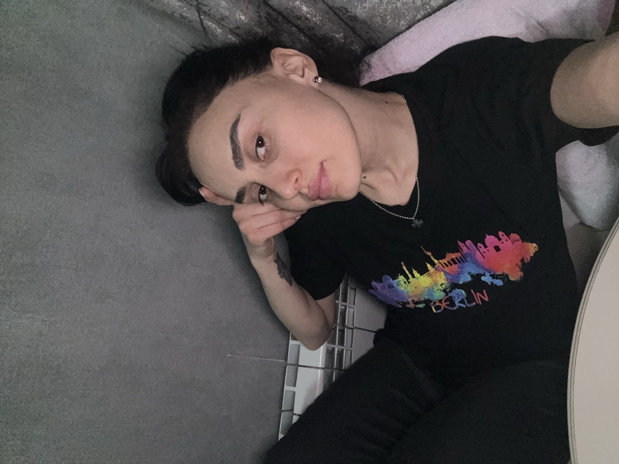 A person with dark hair pulled back is lying down on a grey surface next to a tile wall. They are wearing a black t-shirt with a colorful "Berlin" print and black pants. They have an earring in their left ear and a silver necklace. The person's left hand is resting on their forehead. The background includes a pink towel and a textured grey surface.