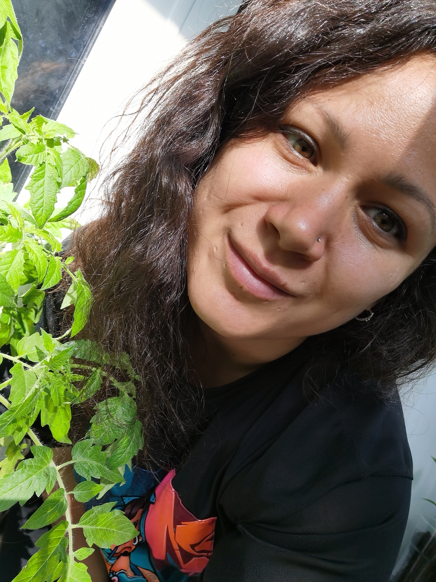 A close-up, high-angle shot depicts a woman indoors next to a plant. The woman has brown curly hair, tan skin, and brown eyes. She is wearing a black shirt with a colorful graphic design on the lower left side. She is smiling slightly, and a nose ring is visible on her left nostril. To the left of the woman is a leafy green plant, possibly a tomato plant, positioned next to what appears to be a window or glass pane. The background is partially obscured by the window and the plant. The location is not discernible from the image.