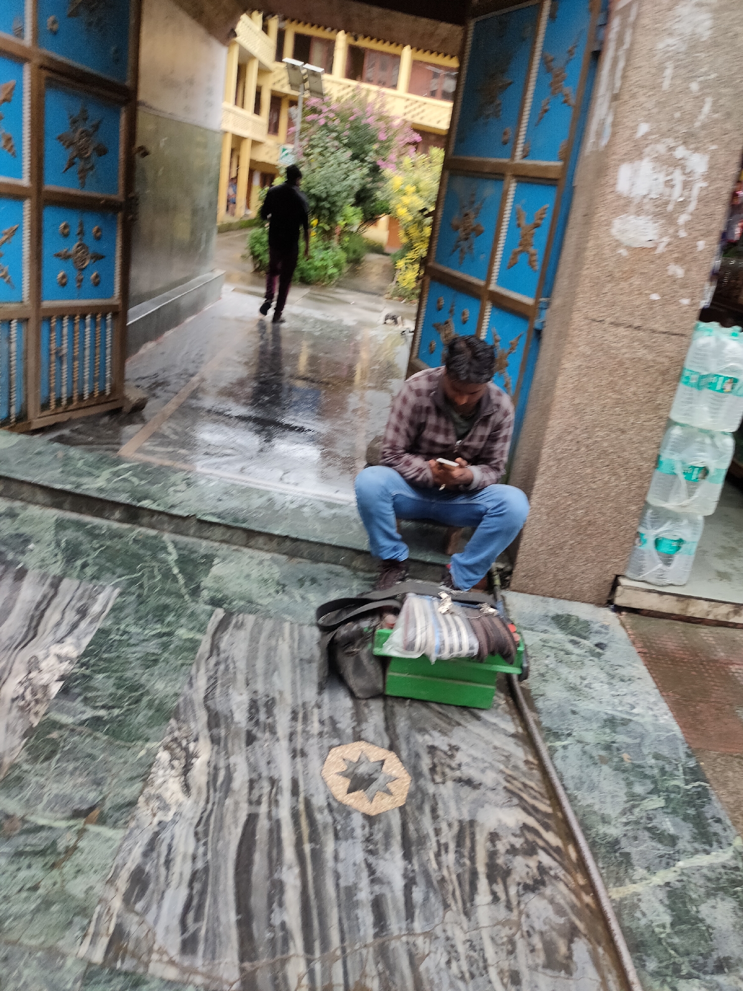 The image depicts a scene outside a building in Manali, India, likely a guesthouse or hotel, given the architecture and the presence of what appears to be a stack of bottled water outside. The ground is wet, suggesting recent rain or a damp environment.
In the foreground, a man is seated on a step, engrossed in his mobile phone. He is wearing a plaid shirt and blue jeans. Beside him is a dark bag and a green rectangular box containing various items, including what looks like folded textiles or bags. The floor is made of polished green and grey marble, with water reflecting the surroundings. A decorative star inlay is visible on the marble.
Further in the background, another person is walking away from the camera, dressed in black. Behind this person is a building with multiple windows and a bright yellow facade. To the left, a large blue wooden door with intricate metalwork stands open, revealing a glimpse of the interior. To the right, a stack of bottled water is visible, some with green labels. The overall atmosphere suggests a slightly humid or rainy day.