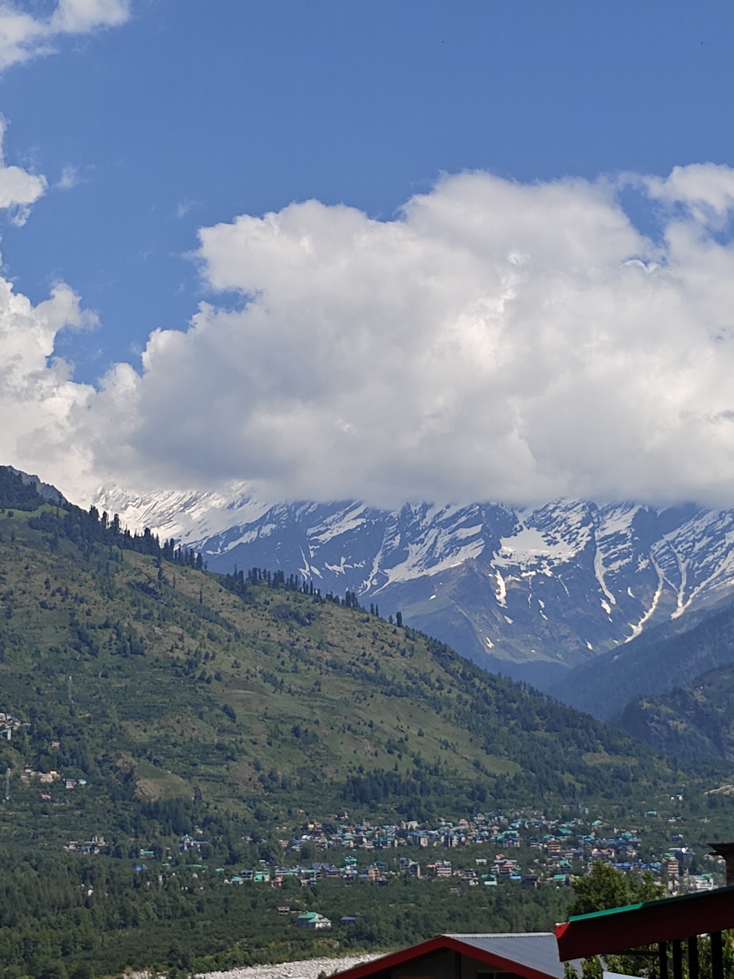 The image depicts a scenic mountain landscape with a town nestled in the valley. In the foreground, lush green hills dotted with trees lead the eye towards a cluster of buildings with colorful roofs, suggesting a town or village. Beyond the town, majestic snow-capped mountains rise sharply against a vibrant blue sky, partially obscured by fluffy white clouds. The overall scene evokes a sense of natural beauty and tranquility, characteristic of a mountainous region. While the image does not contain any people or specific actions, it captures the essence of a picturesque location, possibly Vashisht, India, known for its natural beauty and mountainous terrain.