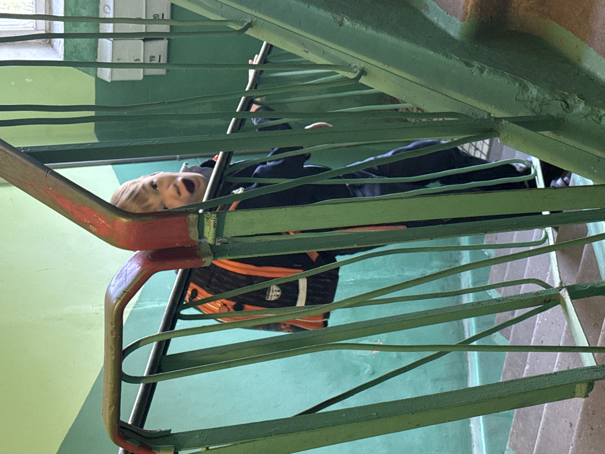 A child with blonde hair and an open mouth is peering through the green metal railing of a stairwell. The child is wearing a dark-colored jacket and carrying a black and orange backpack. The stairwell has green walls, and numbers are visible on the wall behind the railing. The stairs are made of concrete. The image was taken in Dmitrov, Russia.