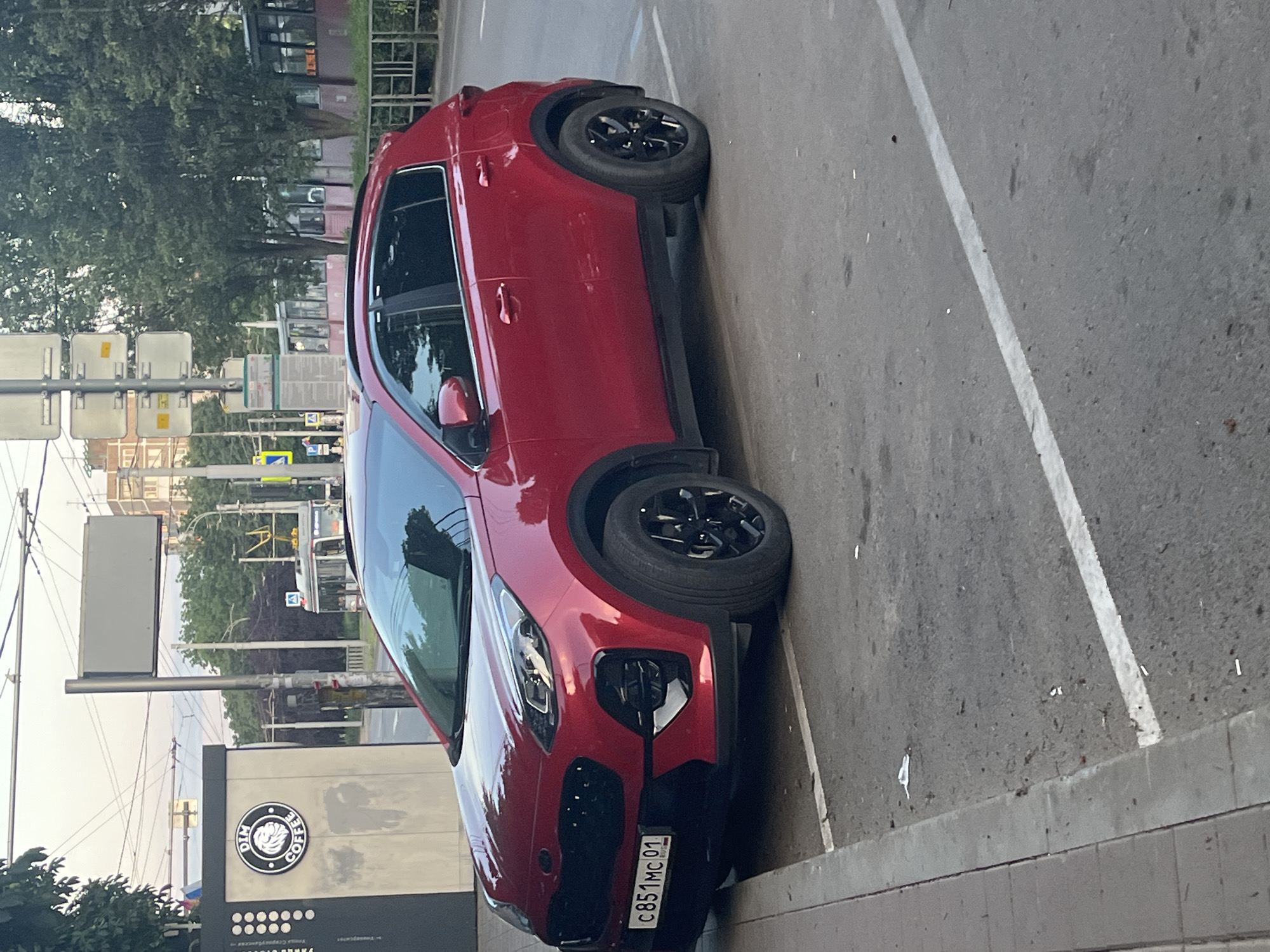A red Kia Sportage is parked on a city street in Krasnodar, Russia. The vehicle is facing left and is positioned in a parking spot indicated by white lines on the asphalt. The car's license plate reads "C851MC 01 RUS". To the left of the car is a sidewalk with a gray curb and a building with a sign that says "DIM COFFEE" featuring a lion's head logo. In the background, trees, buildings, streetlights, and a tram are visible. The sky is overcast.