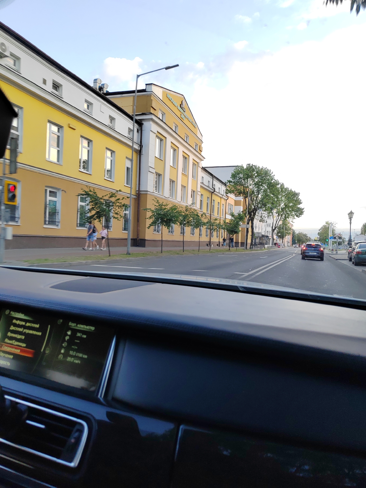 This is a first-person perspective, likely from the driver's seat of a vehicle, looking forward. The scene depicts a city street in Brest, Belarus. In the foreground, the dark dashboard of a car is visible, with a digital display screen showing menus in Russian, including "Settings," "Information Display," "Control Display," "Time/Date," "Language/Units," "Connections," and "Volume." To the right of the display, there is a dark, reflective surface.
The car is driving down a street lined with trees and buildings. On the left, there is a distinct yellow building with white trim and multiple windows. A traffic light with a red signal is visible on the far left. Two people, a man and a woman, are walking on the sidewalk in front of the yellow building.
Further down the street, more buildings are visible, including a white building. The street itself has visible lane markings. Several cars are on the road, with one blue car ahead of the perspective vehicle and other cars parked on the right side of the road. Green trees are planted along the sidewalk. The sky above is mostly clear with some white clouds. The overall impression is of a normal day in a European city.