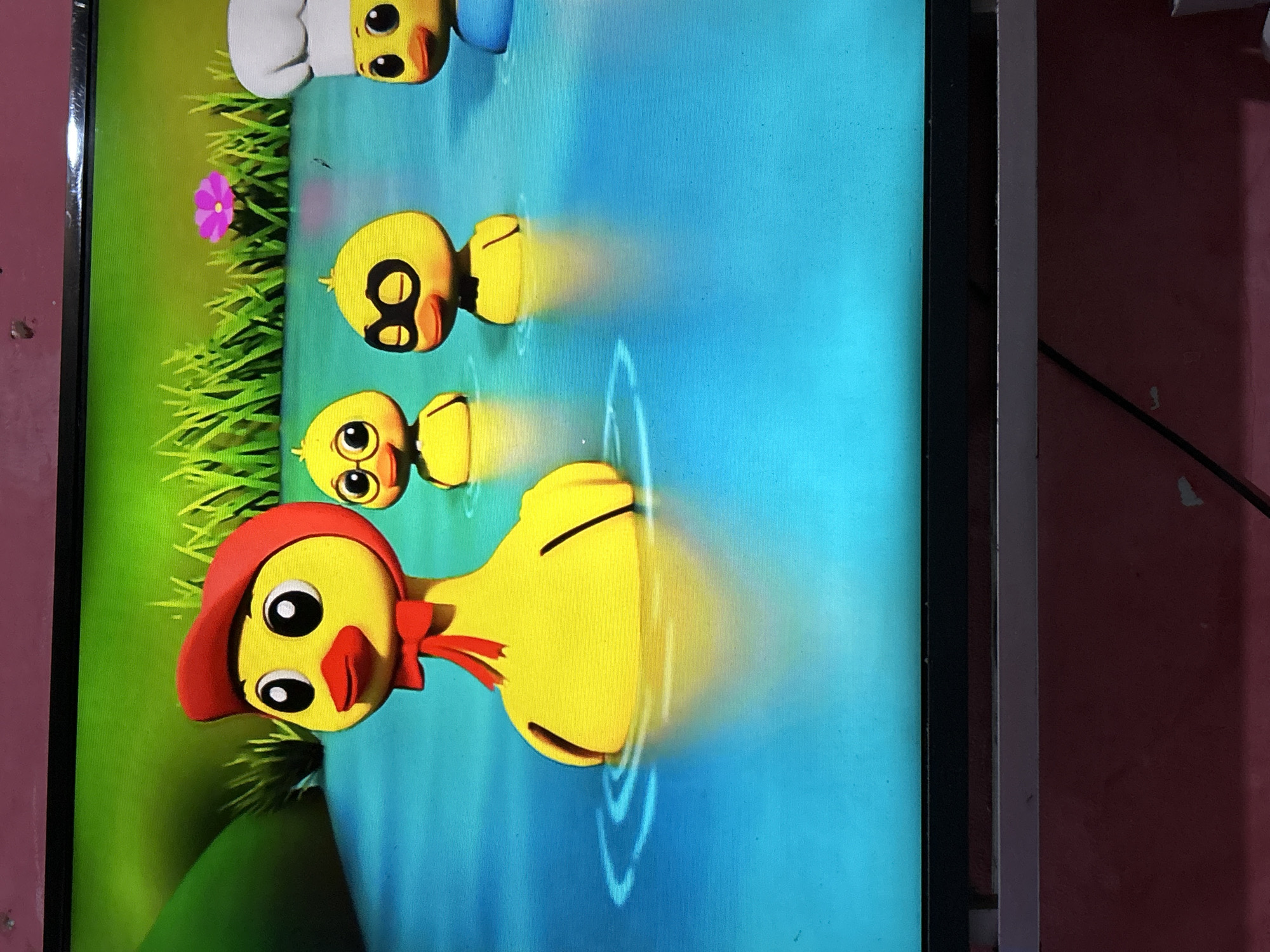 The image shows a television screen displaying a cartoon animation of several yellow ducklings in blue water. The ducklings are depicted with exaggerated features, such as large eyes and simplified bodies. One duckling in the foreground wears a red hat and a red bow. Other ducklings in the background wear different accessories, including glasses and a chef's hat. The background of the animation features green grass and a pink flower. The television is placed on a stand, which is against a red wall. The scene does not depict any elements that would allow for location determination in Depok, Indonesia.