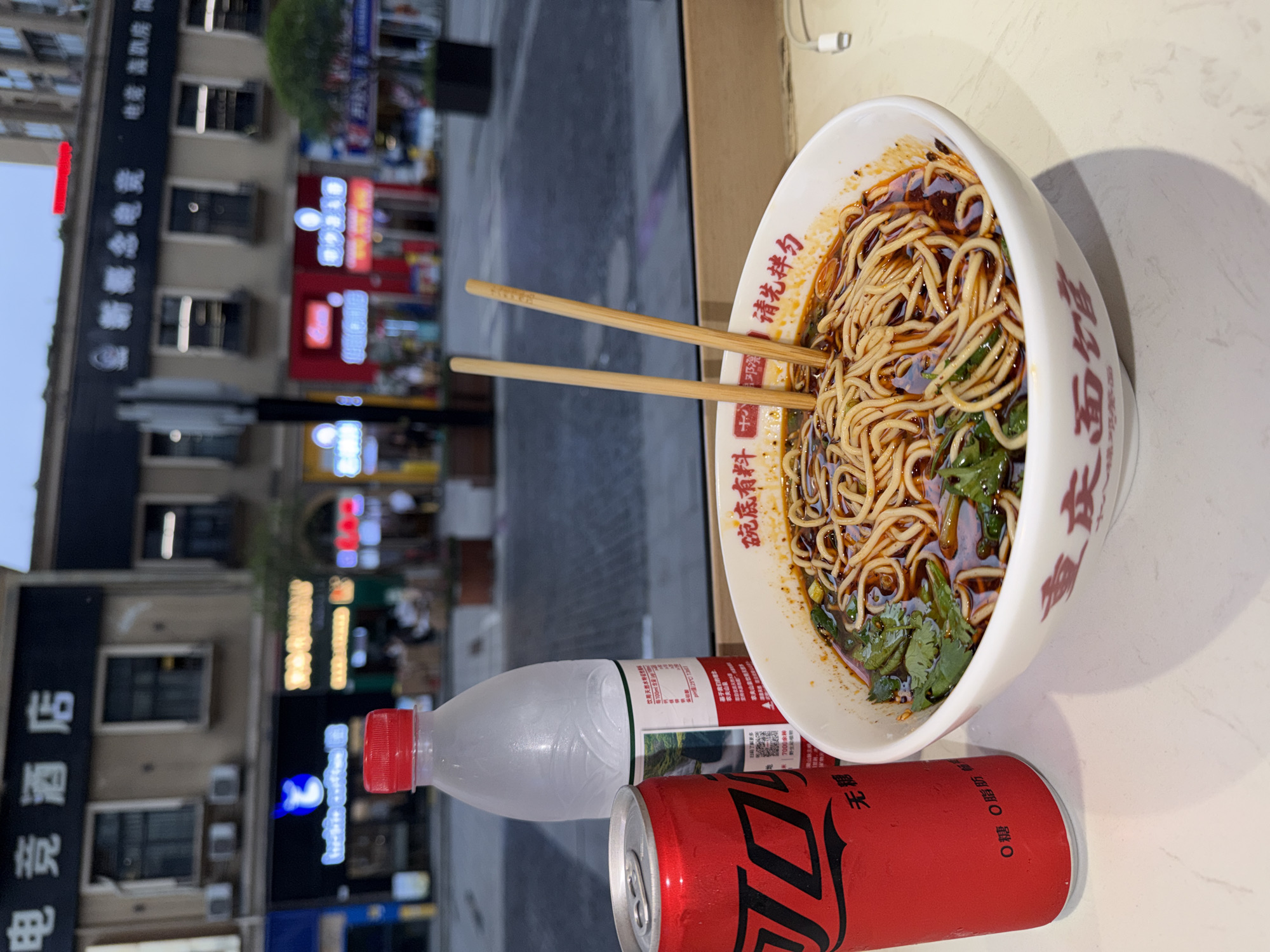 The image presents a close-up of a meal set against the backdrop of an urban street scene at night, located in Chongqing High-tech Zone, China.
In the foreground, a white bowl with red Chinese characters holds a serving of noodles in a rich, reddish chili oil sauce, garnished with green herbs, likely cilantro. A pair of wooden chopsticks rests across the bowl. Text visible on the bowl includes "碗底有料" (Ingredients at the bottom of the bowl), "请先拌匀" (Please mix well first), and "重庆小面" (Chongqing noodles), strongly suggesting a local specialty.
Beside the bowl are two beverages. A clear plastic bottle of Nongfu Spring water, featuring a red cap and a label showing "农夫山泉" (Nongfu Spring) and "饮用天然水" (Drinking Natural Water), is positioned next to a red can of Coca-Cola Zero. The can's text reads "无糖" (Sugar-free) and "0糖 0脂肪" (0 sugar 0 fat).
The background, blurred but discernible, shows multiple-story buildings lining a street, illuminated by various neon and LED signs. The time of day is clearly night due to the prominent artificial lighting. Notable signs on the buildings include "电竞酒店" (E-sports Hotel) and "新概念电竞" (New Concept E-sports), indicating the presence of gaming or technology-related establishments in the vicinity. The scene captures a casual dining experience in a vibrant, modern city environment.