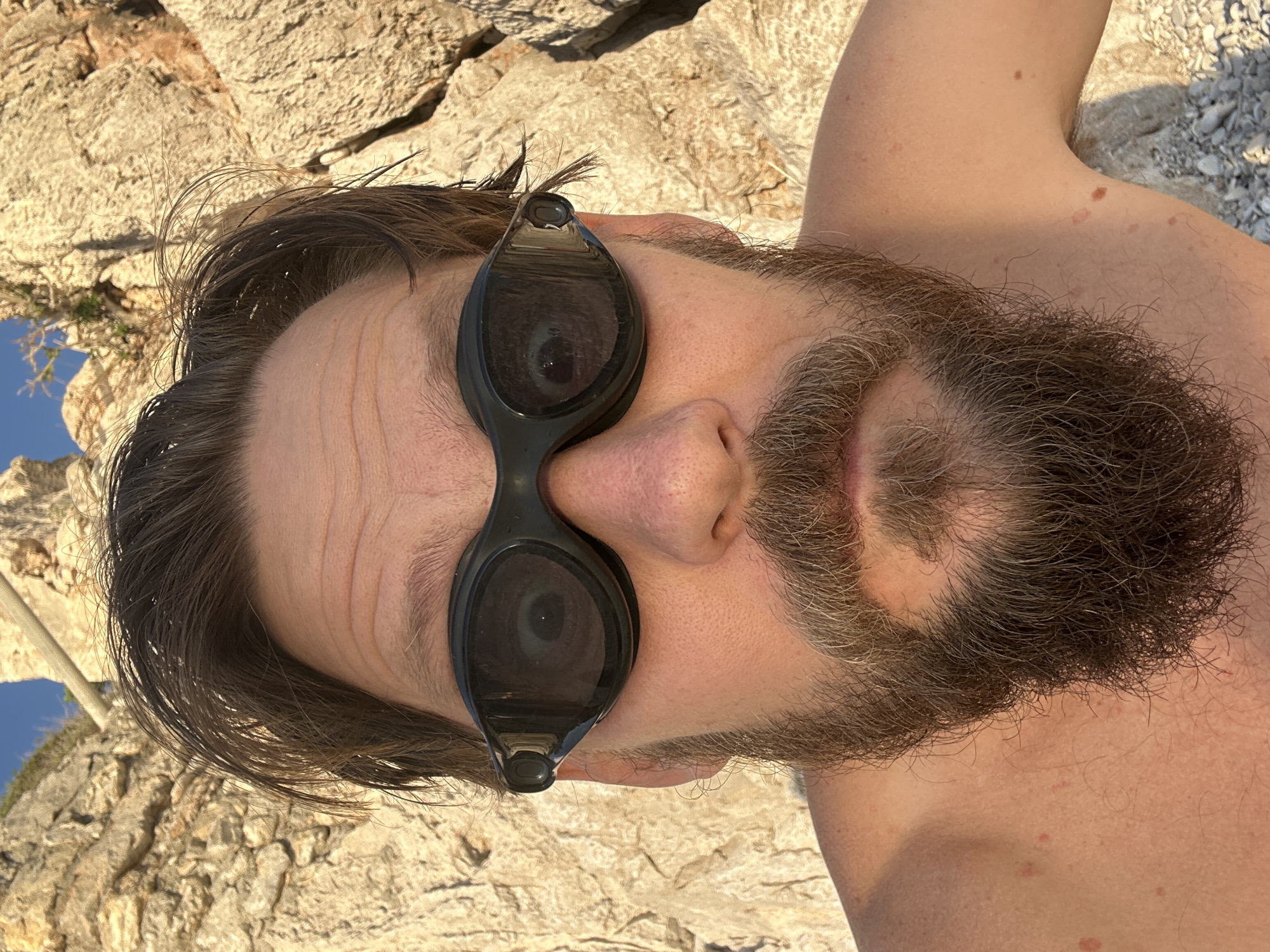 This close-up image presents a person, likely male, lying down in an outdoor, sunlit setting. The photo is rotated 90 degrees counter-clockwise, showing the person's head on the left and upper torso extending to the right. The subject has dark brown hair, appearing slightly disheveled, and a prominent, full dark brown beard with streaks of grey. They are wearing black-framed swimming goggles with dark, reflective lenses that obscure the eyes. The forehead shows some horizontal lines. The person's bare shoulder and upper arm are visible on the right side of the frame, exhibiting several moles and some subtle reddish patches on the skin.
The background is dominated by light-colored, textured rocks, suggesting a rocky coastline or beach area. The rocks are brightly illuminated by the sun, creating clear shadows and highlighting their rugged surface. A small section of clear blue sky is visible in the upper left corner, along with a glimpse of what might be distant land or foliage. The overall atmosphere is bright and warm, indicative of sunny weather. The scene suggests a moment of relaxation, possibly after a swim or while sunbathing on the rocky shores of Fethiye, Turkey. There is no visible text in the image.