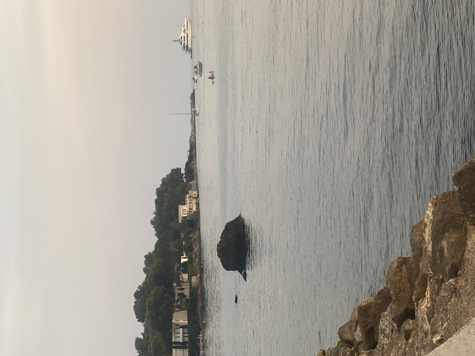 The image depicts a serene seascape off the coast of Antibes, France. In the foreground, a rocky shoreline with tan-colored stones lines the bottom left of the frame. The sea, a calm expanse of blue-gray water with subtle ripples, dominates the majority of the image. A large, dark, irregular rock formation emerges from the water near the center of the frame, with two smaller rocks visible to its left.
In the middle ground, a tree-lined coastline stretches across the horizon. Several white, modern buildings with flat roofs are nestled amongst the trees. Small groups of people are visible on a paved walkway or beach area along the coastline, some appearing to be sunbathing or walking.
Further out in the water, several boats are present. A large, white superyacht with multiple decks and a tall superstructure is positioned on the right side of the frame. To its left, a smaller white boat is moving. In the distance, a white sailboat with its mast visible is anchored, and another small boat is visible further left. Two people are on a paddleboard in the middle distance. The sky above is a hazy, pale gray, suggesting either early morning or late afternoon light. The overall impression is one of a peaceful coastal scene on a clear day.