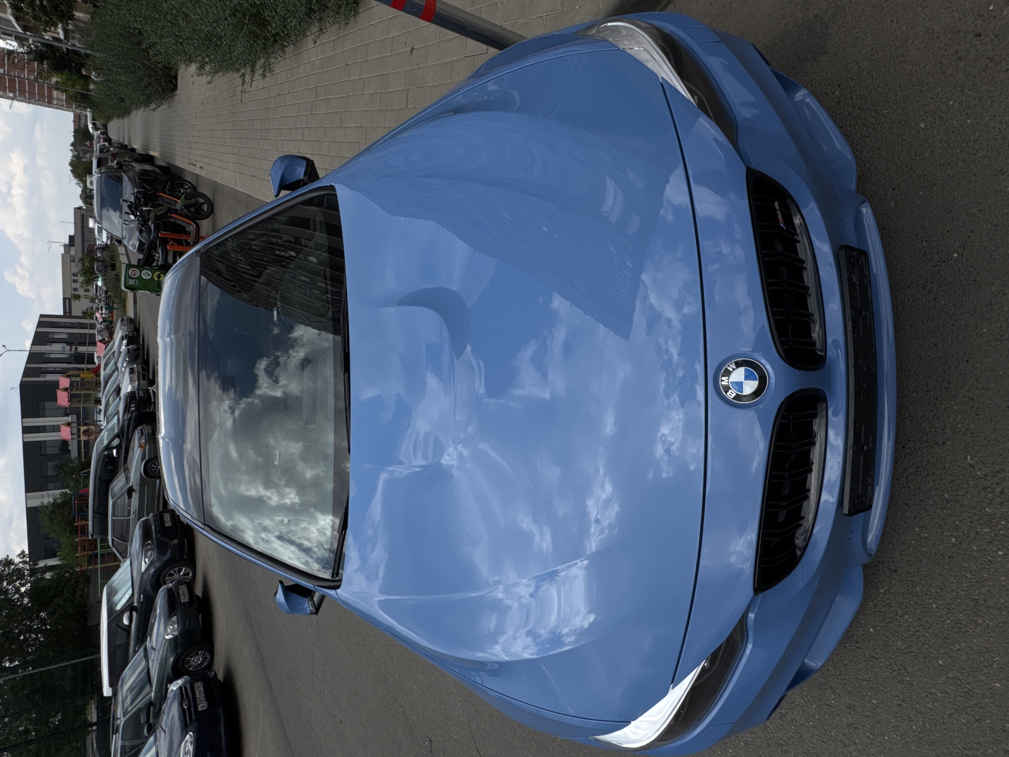 A blue BMW M4 coupe is parked on a paved surface, facing forward. The car is the primary subject, positioned centrally in the frame. The hood of the car reflects a cloudy sky. The front grille and BMW emblem are visible. In the background, other vehicles are parked, including SUVs and sedans, on a street adjacent to a sidewalk. Buildings and trees are visible further in the distance. The scene appears to be in an urban or suburban setting.