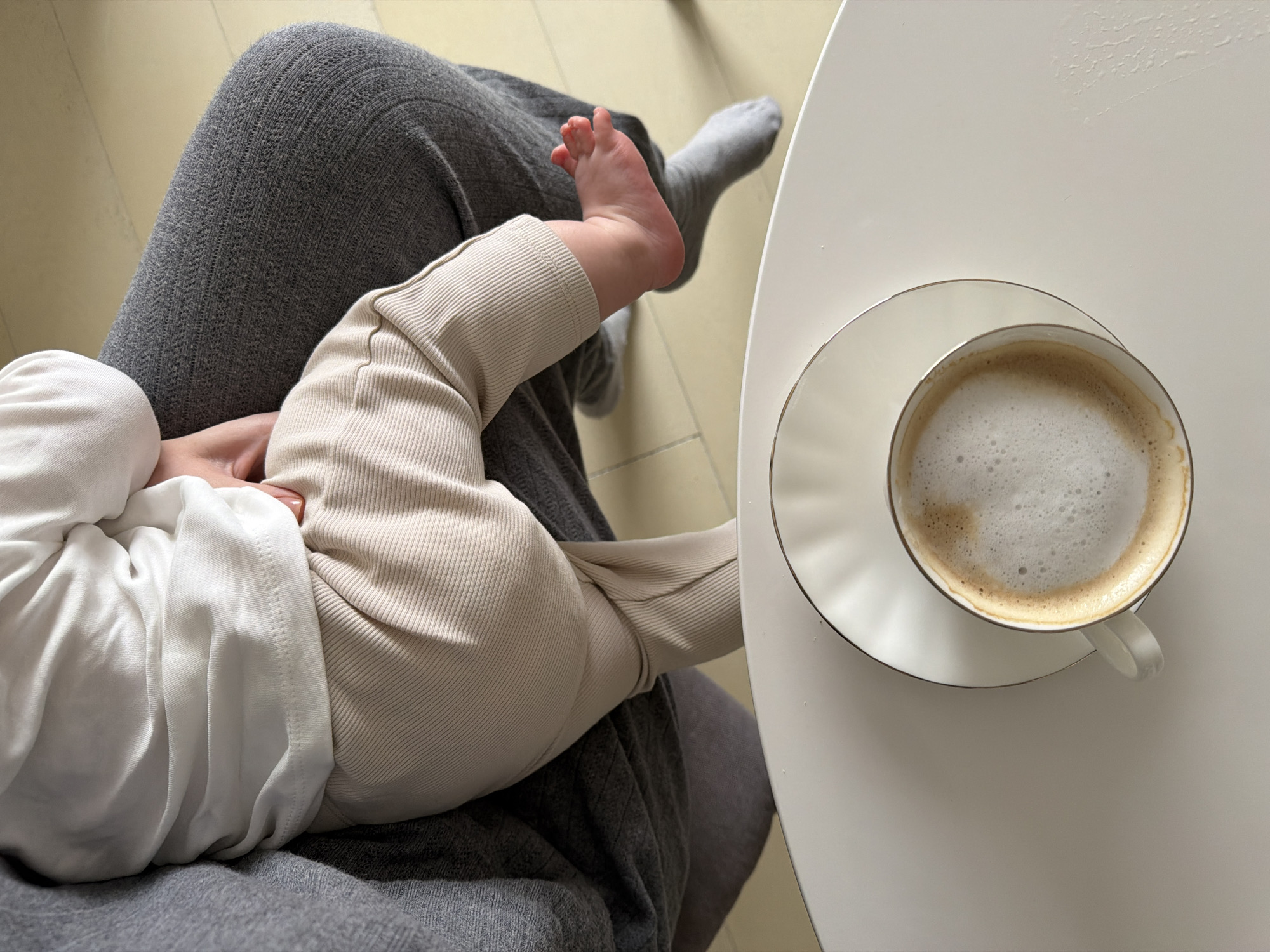 A top-down shot shows a person holding a baby while sitting at a table with a cup of coffee. The person is wearing gray pants. The baby is wearing a white shirt and beige pants. The coffee cup is white and on a white saucer, placed on a white table. The floor is pale yellow. The exact location in Khimki, Russia, cannot be determined from this image.