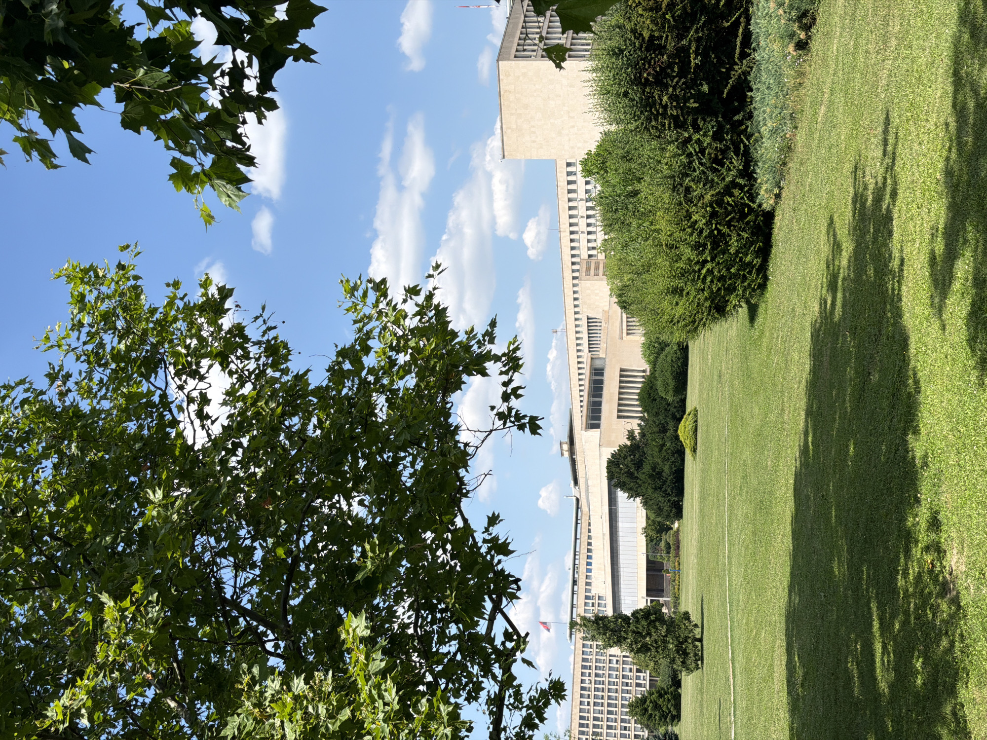 The image depicts a wide lawn with a building in the background, viewed from a park-like setting. The foreground is dominated by lush green grass, partially shaded by tree branches. Several mature trees and shrubs are scattered across the lawn, with some denser bushes framing the right side of the image.
In the middle ground, a large, sprawling building with a light-colored facade and numerous windows is visible. A flag pole with a flag is positioned to the left of the building. The building appears to be a modern or modernist architectural style, possibly an institutional or governmental complex.
The background consists of a clear blue sky with scattered white clouds. The overall impression is one of a sunny day in a well-maintained urban park or campus. Based on the architectural style of the building and the surrounding landscape, this scene is likely located in Belgrade, Serbia, possibly depicting the Palace of Serbia, which is known for its grand architecture and expansive grounds. There are no discernible people or specific actions taking place in the image.