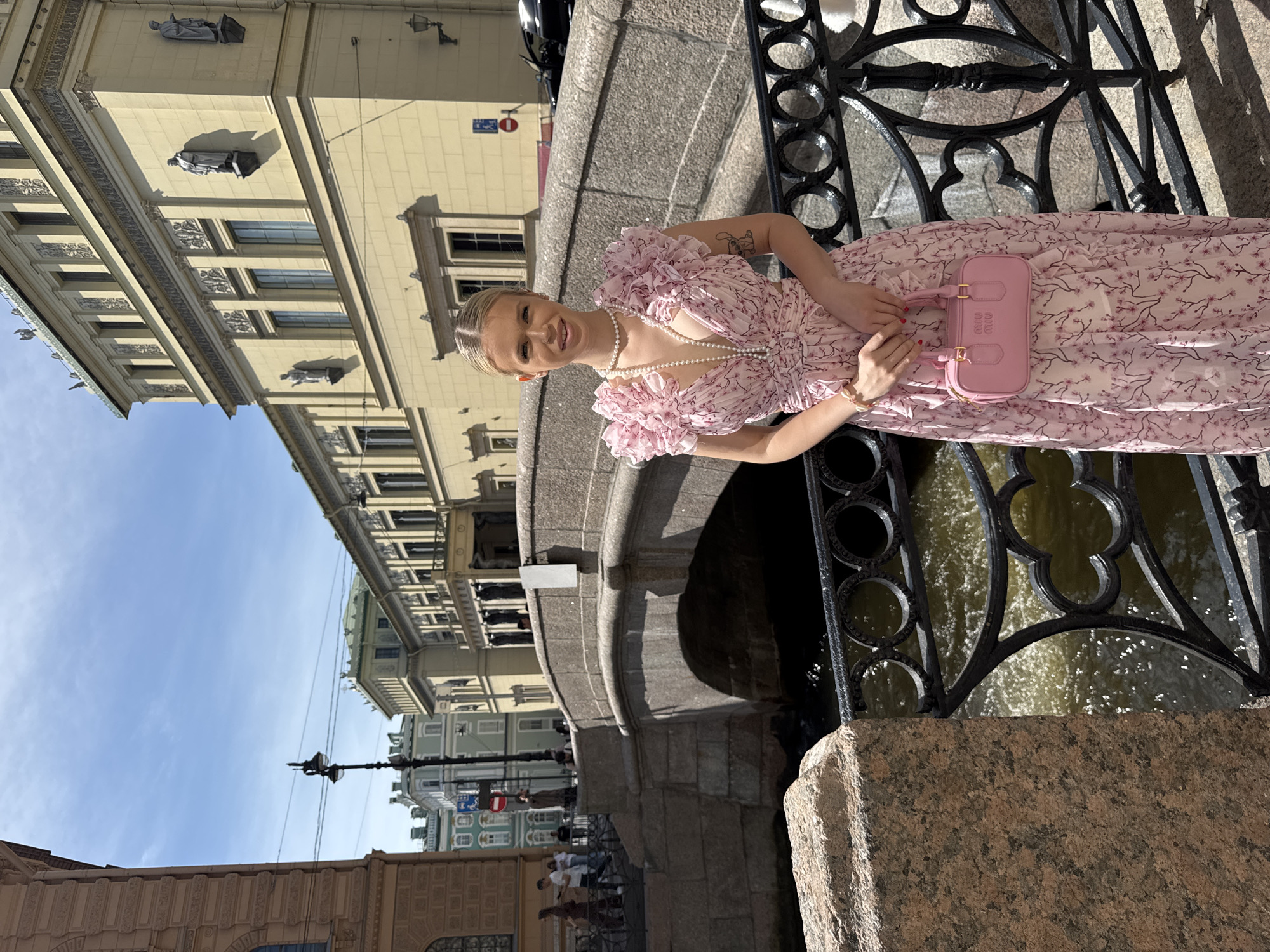 In this portrait, a young woman with blonde hair styled in a bun poses beside a decorative black iron fence overlooking a canal. She is wearing a light pink floral dress with ruffled sleeves and a pearl necklace. In her hands, she holds a small pink handbag with "MIU MIU" embossed on it. Her expression is one of a slight smile, suggesting a pleasant mood. The background reveals the architecture of Saint Petersburg, with ornate building facades and statues visible. The canal below has water reflecting the sunlight, indicating a bright day. The presence of classical European architecture, along with the canal setting, strongly suggests a location in Saint Petersburg, Russia. The time of day appears to be daytime, given the strong sunlight casting shadows.
