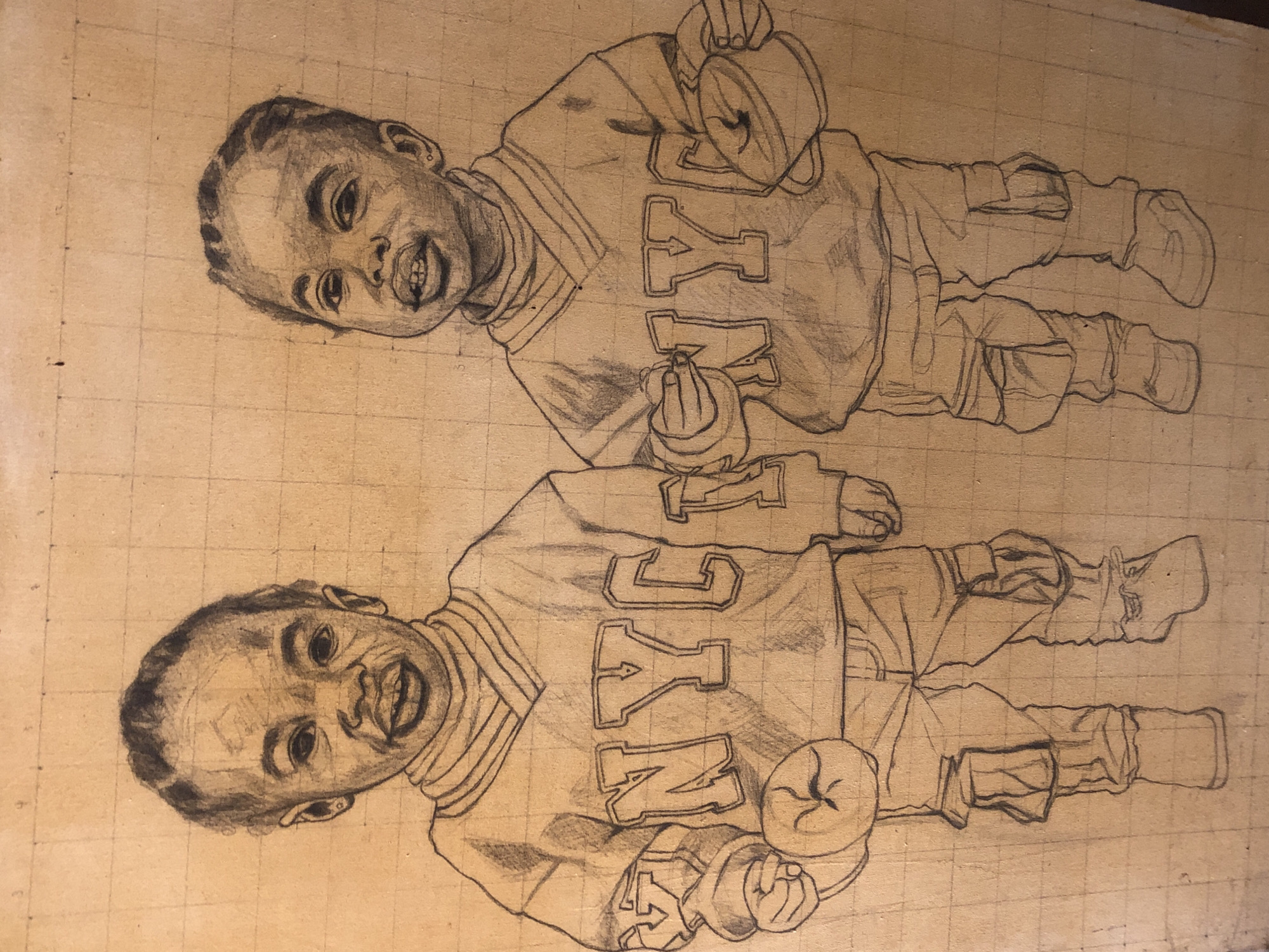 Two identical pencil drawings on a tan surface, possibly wood, overlaid with a grid pattern. The drawings depict a young child in a jersey with the letters "NYC" on the front. The child holds a cup or container in one hand and a small object in the other. The subject is wearing pants and shoes with details like straps and pockets. Both drawings are oriented in the same direction with the upper drawing slightly rotated. The exact location in Ado, Nigeria cannot be determined from the image.