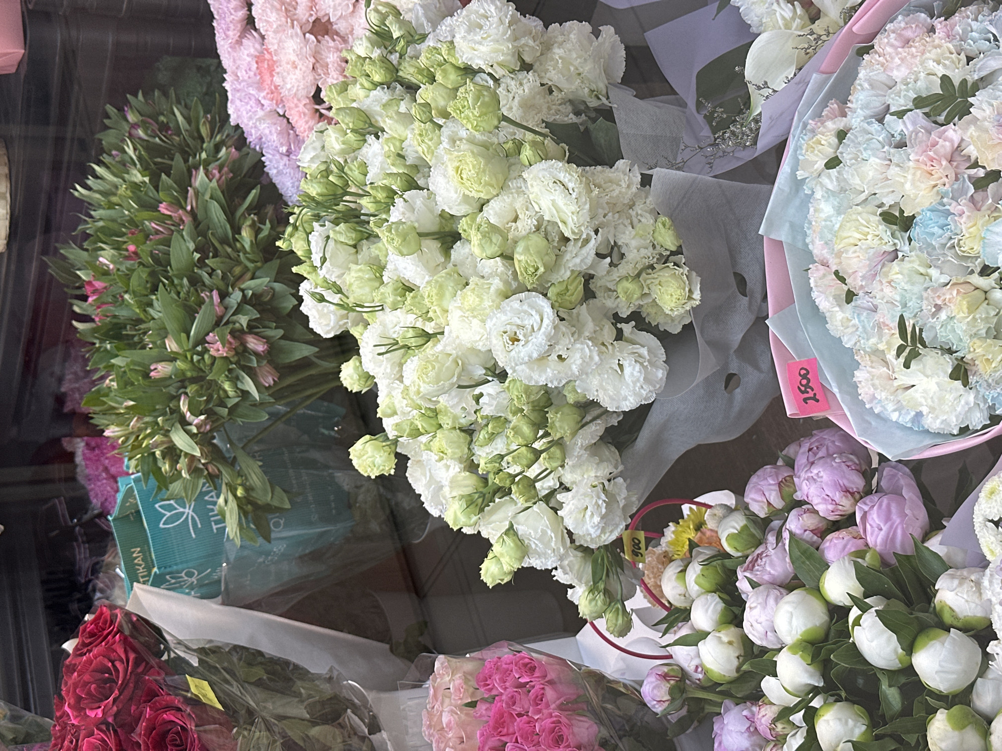 A variety of fresh flower bouquets are displayed in a shop window, with some featuring price tags. The bouquets include red roses, pink roses, white and green eustomas, pink carnations, peonies in pink and white, and a bouquet of multi-colored carnations in pastel shades. There are also two teal-colored gift bags with a white lotus-like logo visible behind some of the flowers. The scene appears to be a flower shop in Novoaltaysk, Russia. There are no people visible in the image.