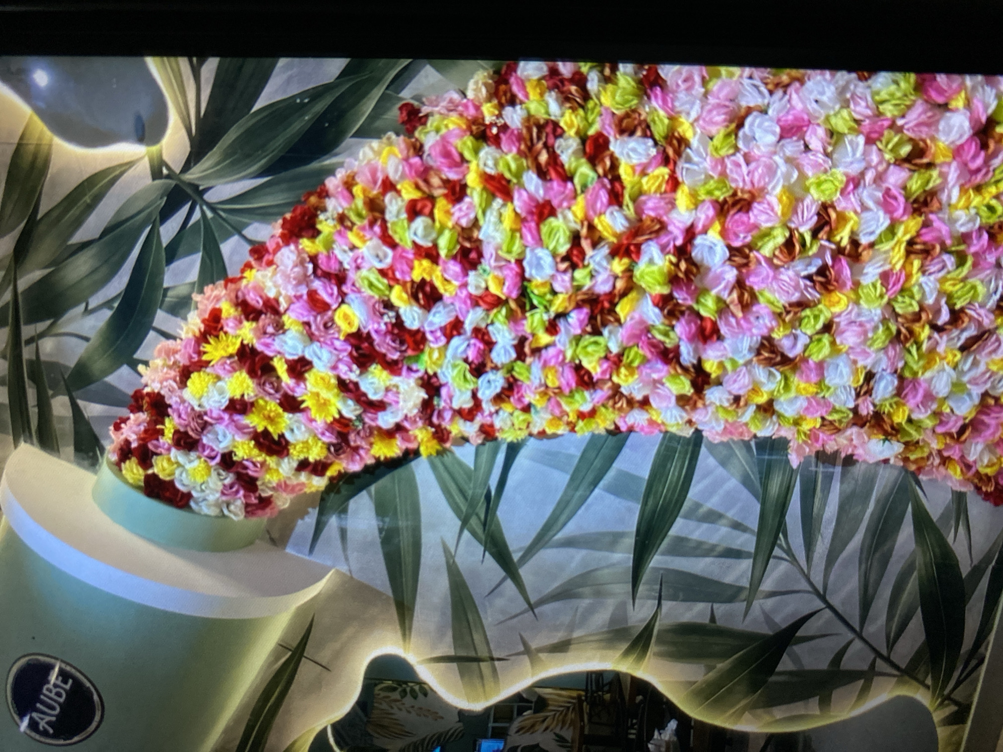The image depicts a close-up of a decorative installation featuring a large, light green cylindrical object, reminiscent of a tube of paint, tilted and seemingly dispensing a cascade of colorful, small fabric flowers. The flowers are a vibrant mix of pink, yellow, red, white, and green, creating a textured, overflowing effect. Behind the floral cascade, a stylized, pale silhouette of a figure or torso with a prominent curve is visible, superimposed on a background of large, green palm leaves. The overall impression is one of a whimsical and artistic display, likely found in a public space or shop. There is a circular emblem on the cylindrical object with the word "AUBE" in white text. The lighting suggests an indoor setting, possibly a retail store or exhibition space, with soft illumination highlighting the details of the installation. The city of Ta', Indonesia is not depicted or mentioned in the image.