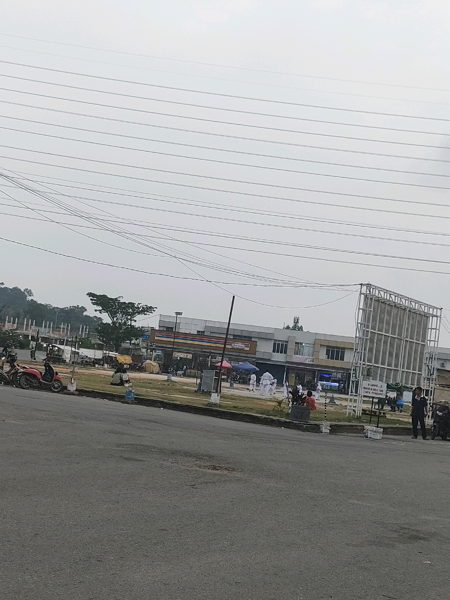 The image shows a street scene in Batam, Indonesia, likely near a commercial area. In the foreground, a gray asphalt road is visible with some patches and cracks. To the left of the road, there is a grassy area with several red and black scooters parked. In the background, a strip of buildings is situated, including what appears to be a convenience store with a prominent red, yellow, and blue striped awning. Further to the right, a large, empty billboard structure is present. Several people are scattered throughout the scene, including individuals in martial arts uniforms on the grassy area, a man in a dark suit standing near the billboard, and others milling about in front of the buildings. Overhead, a complex network of power lines crisscrosses the sky, which is overcast. The environment suggests a somewhat busy, everyday scene in an Indonesian urban setting.