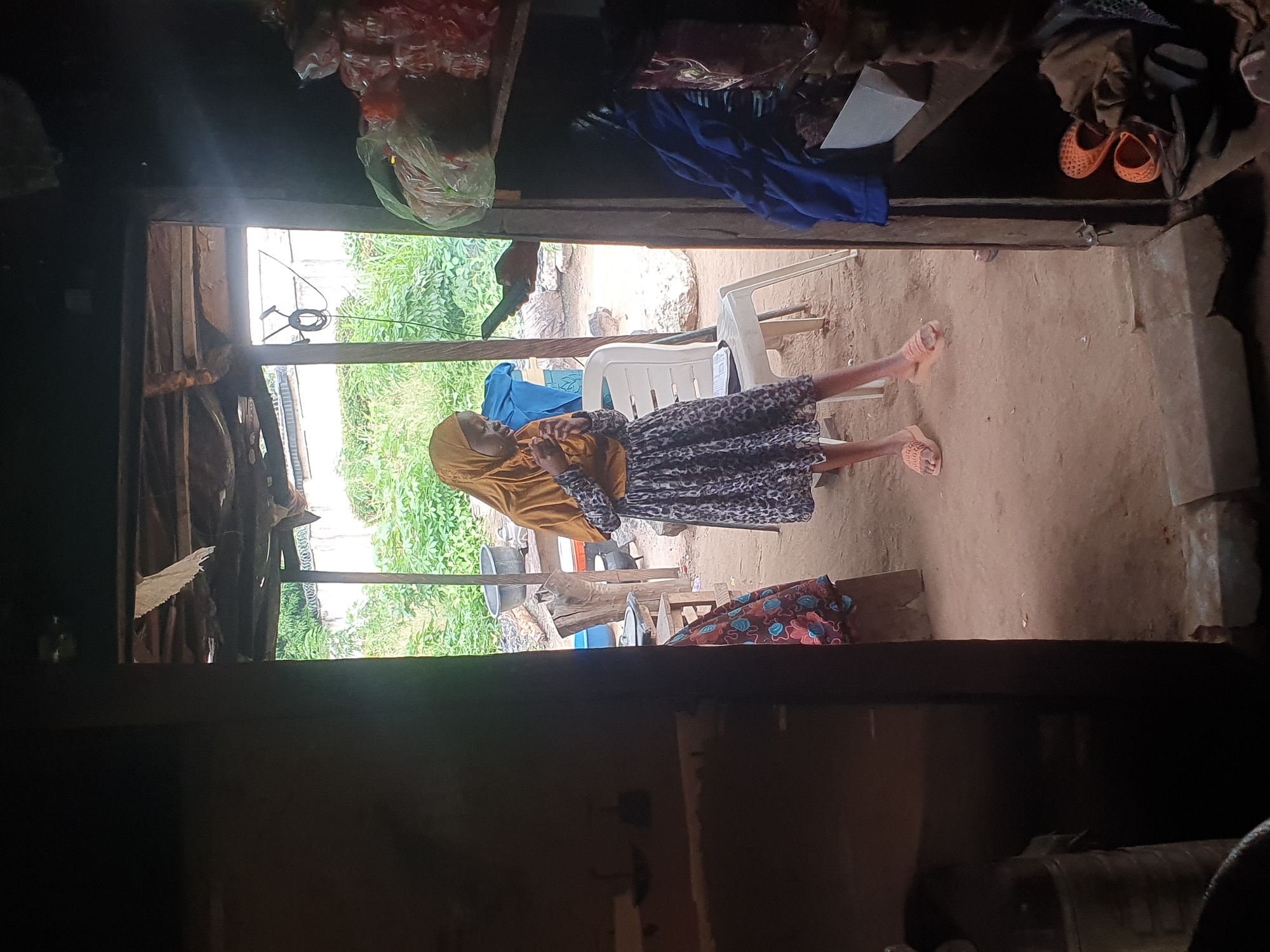 In Lokoja, Nigeria, a young girl wearing a yellow hijab and a patterned dress stands in a doorway. She is holding what appears to be food in her hands. To her right, a white plastic chair is visible, and further back, an assortment of cooking pots and other items are scattered in a yard. The doorway opens into a rustic interior, with shelves stocked with various goods on the right side. The ground is dirt, and the ceiling appears to be made of corrugated metal and wood. The overall scene depicts a humble, outdoor kitchen or living space.