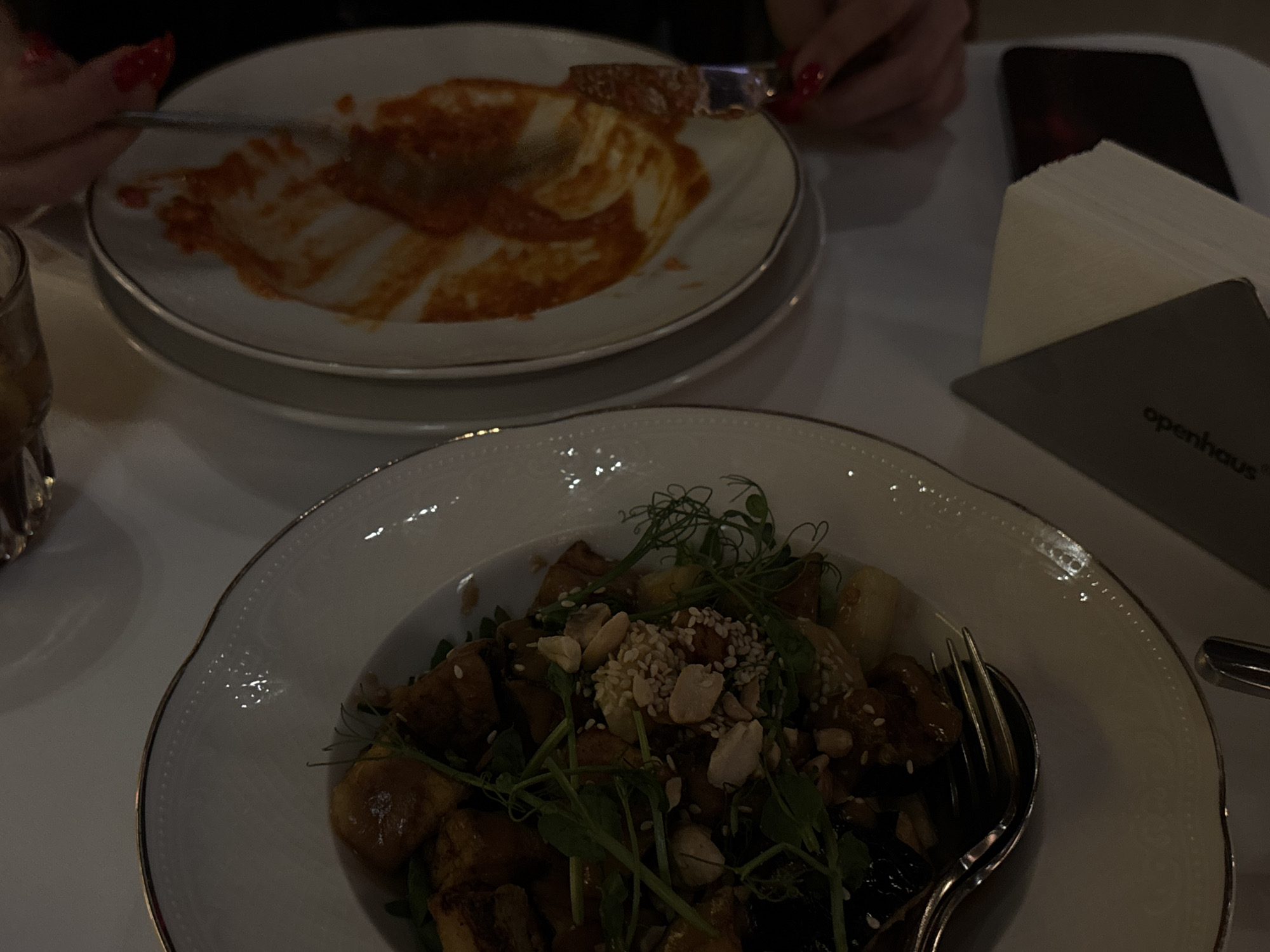 A table setting shows two plates of food, silverware, a glass, a phone, and napkins. The upper plate contains the remnants of a red-sauce dish, partially consumed. The lower plate presents a salad with greens, nuts, and what appear to be mushrooms or roasted vegetables. A fork and spoon are arranged on the plate. The scene is possibly from a restaurant in Yerevan, Armenia, indicated by the "openhaus" detail on the napkin holder. Two hands with red nail polish are visible. One hand is holding a knife, and the other hand is holding a fork.