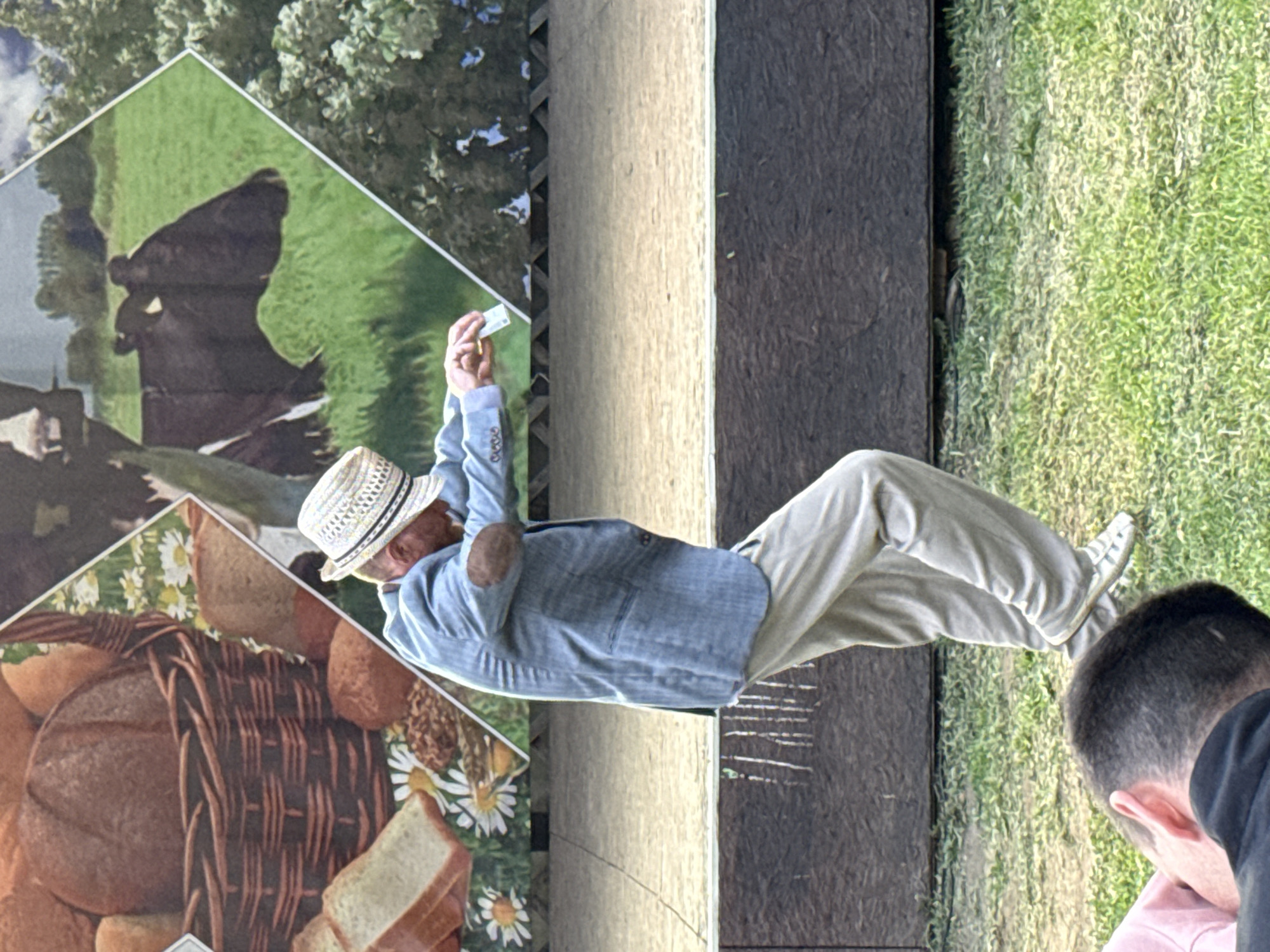 A man in a light blue blazer, light-colored pants, and a straw hat with a black band is dancing on a grassy area in front of a stage. The man is leaning forward with his arms extended, holding a small white object in his right hand. He is wearing brown elbow patches on his blazer. Behind him is a backdrop depicting a basket of bread, a cow in a field, and trees. The ground is covered in green grass and there is a wooden structure in front of the stage. In the foreground, the back of a person's head wearing a black shirt is visible, partially obscured by a pink garment. The image appears to have been taken outdoors in a location that could be a park or festival. The image does not contain any recognizable landmarks or elements that would allow for precise identification of the city of Кукуи, Russia, or any specific location within it.