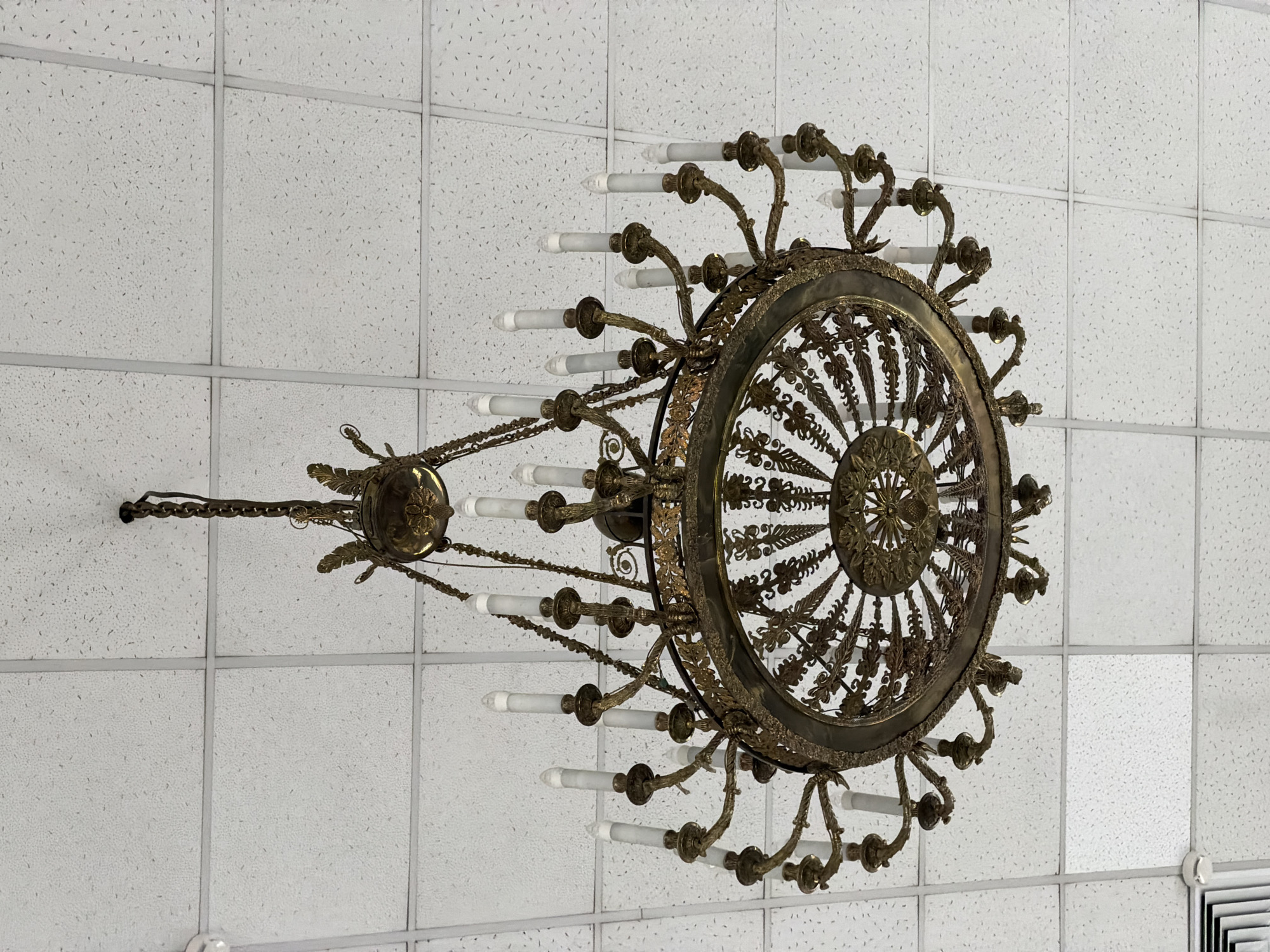The image captures a large, ornate chandelier suspended from a white acoustical tile ceiling. The chandelier, likely made of brass or bronze, exhibits intricate scrollwork, leaf motifs, and a detailed central circular design. Numerous arms extend from its circumference, each supporting a candle-shaped electric light bulb, many of which appear unlit or dim. The chandelier is connected to the ceiling by a sturdy chain, visible towards the top left of the frame.
The ceiling itself is composed of standard white, rectangular acoustical tiles with a speckled texture, held within a visible gray grid system. The perspective is from below, looking upwards, highlighting the contrast between the elaborate, traditional design of the chandelier and the functional, modern appearance of the suspended ceiling tiles. The chandelier is positioned somewhat off-center and at an angle within the frame. There is no discernible activity occurring, nor are any people visible. The setting is an interior space in a building located in Izhevsk, Russia. No visible text is present in the image.