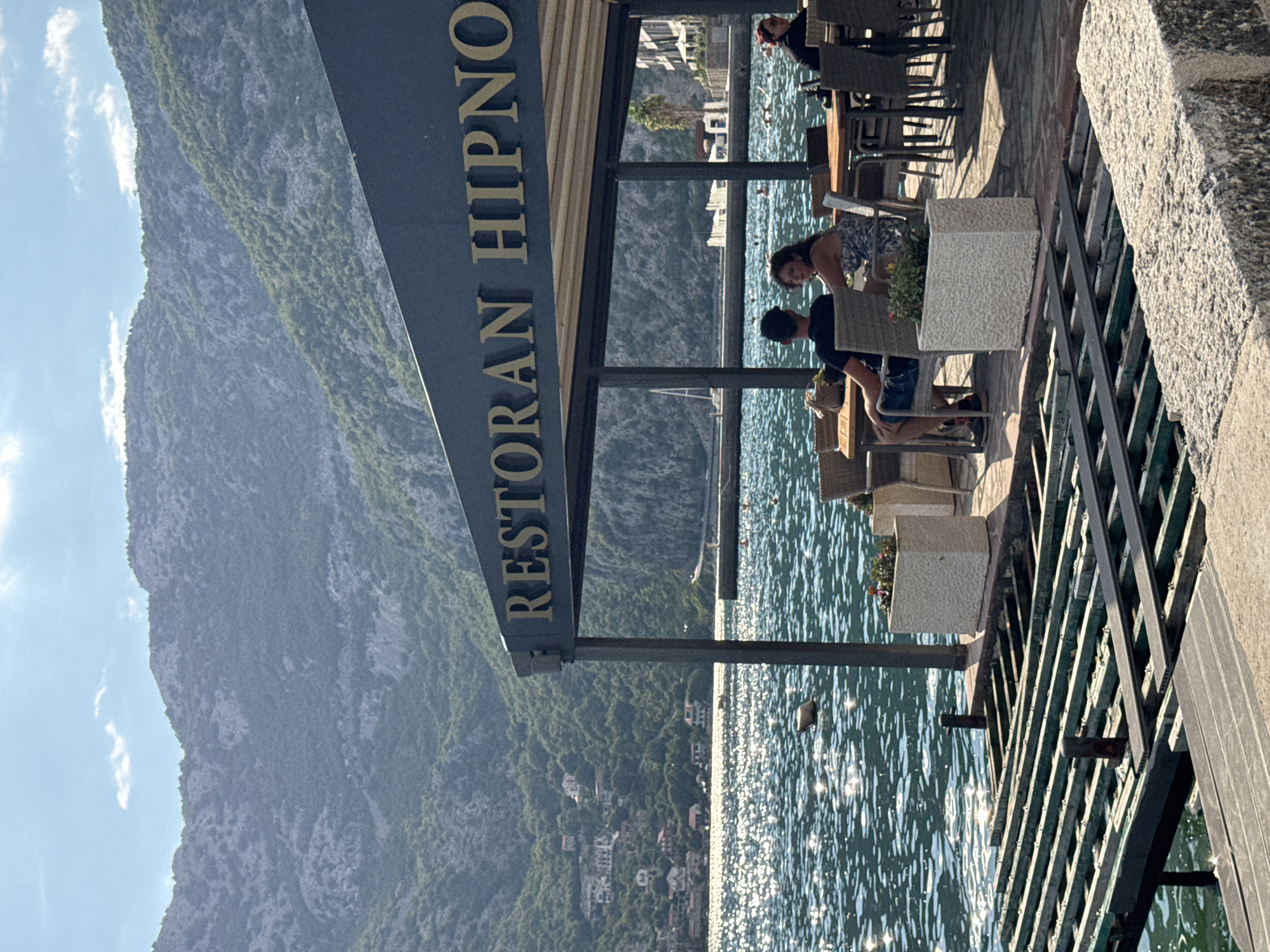 The image depicts a waterfront restaurant called "RESTORAN HIPNO" in Risan, Montenegro. The restaurant has an outdoor seating area with tables and chairs under a dark awning. Several patrons are seated at the tables, with two individuals visible in the foreground. In the background, a body of water, likely a bay or sea, is illuminated by sunlight, creating a shimmering effect. Mountains covered in lush vegetation rise steeply behind the water, with houses scattered along their slopes. A boat with a white sail is visible on the water. In the foreground, there is a wooden pier structure. The overall scene conveys a relaxed, sunny, and picturesque atmosphere typical of a coastal resort.