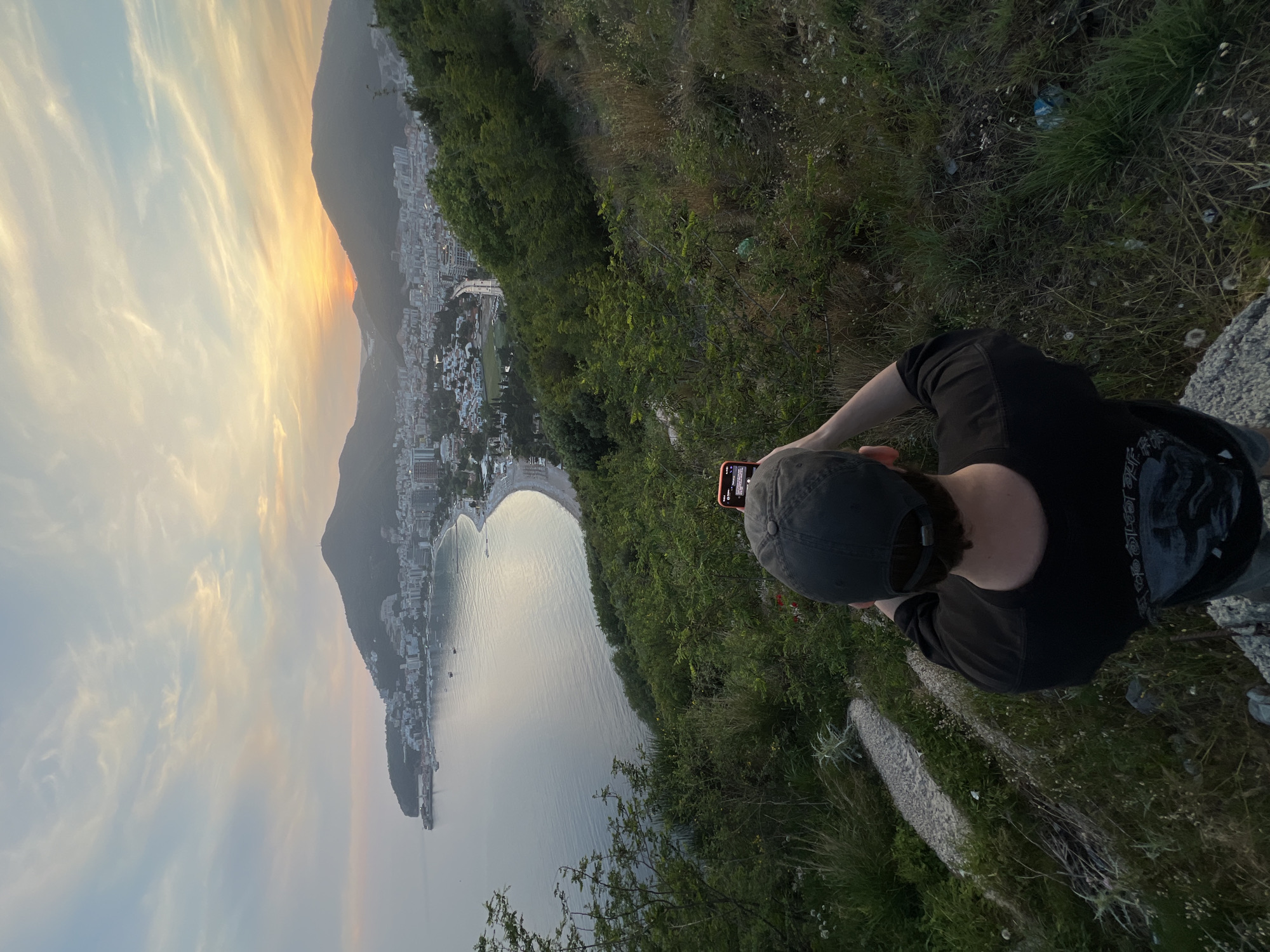 A person is holding a phone and taking a photo of a coastal cityscape and landscape at dusk or dawn. The city buildings are situated between a mountain and a crescent-shaped beach. A body of water is in the foreground. The scene is viewed from a high vantage point with some vegetation. This is Budva, Montenegro.