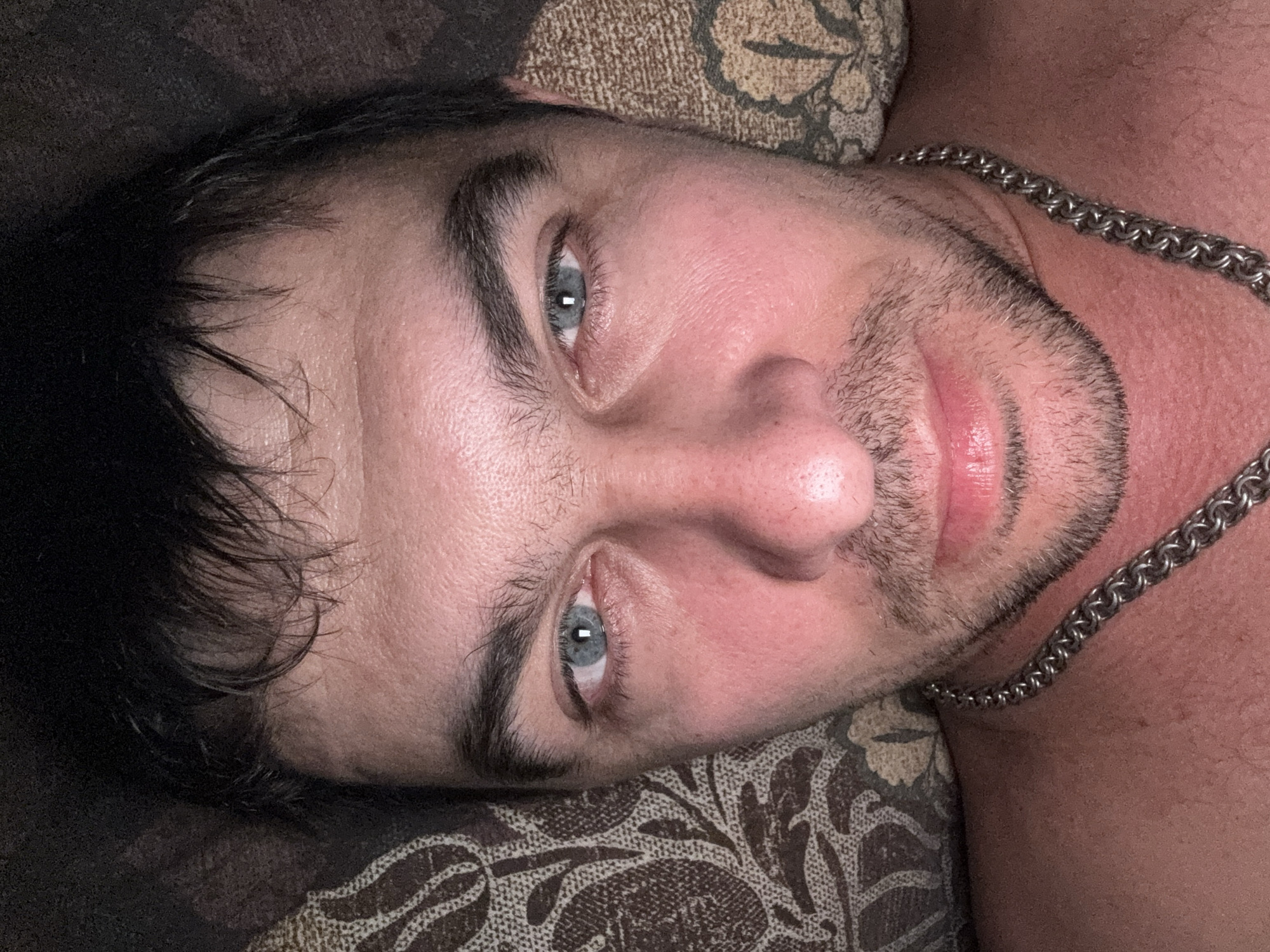 A close-up, head-on, selfie-style shot features a man with dark hair, light blue eyes, and a light beard. He is wearing a silver chain around his neck. The background consists of a patterned surface, possibly a pillow or blanket. The lighting is dim, creating a warm glow on his skin. There is no discernible location within the city of Shakhty, Russia, depicted in this image; it appears to be an indoor scene.