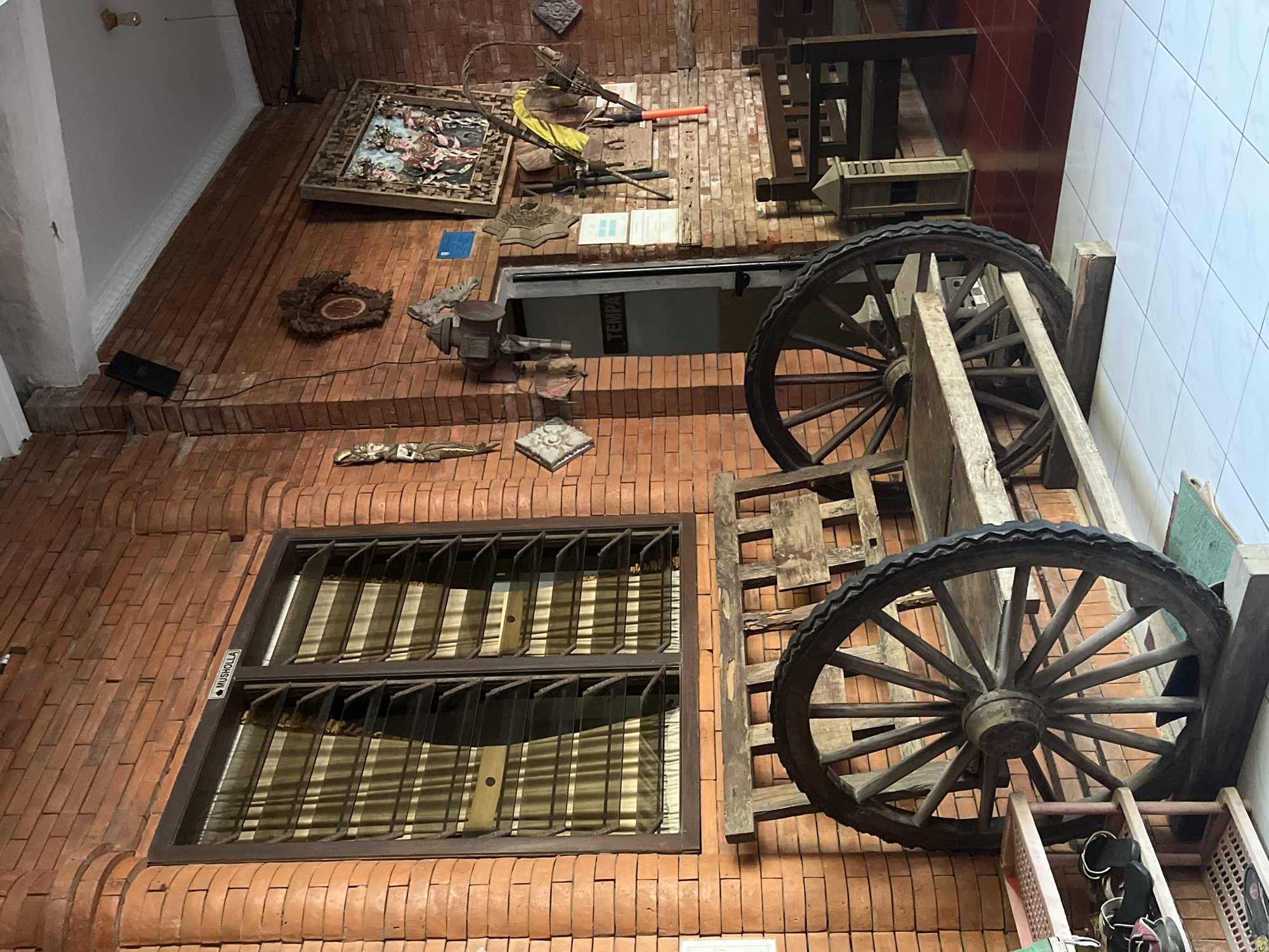 The image depicts the interior of a building with exposed brick walls. In the foreground, a wooden bench with large, old-fashioned wagon wheels on either side is the central focus. To the left, a multi-tiered shoe rack holds several pairs of footwear. Behind the bench, a window with horizontal blinds is visible, and a sign above it reads "MUSHOLLA" with a small crescent moon icon.
The background reveals more of the brick wall adorned with various decorative items, including framed artwork, wooden carvings, and what appear to be traditional implements. A door with a sign reading "TEMPA" is slightly ajar. Further back, more wooden seating arrangements and flooring are visible.
This scene is located at Museum Kartini in Jepara, Indonesia. The museum, dedicated to the national heroine Raden Adjeng Kartini, is known for preserving artifacts and showcasing the heritage of Jepara. The architectural style and decorative elements in the image are consistent with the traditional Indonesian aesthetic often found in such historical sites. The overall impression is one of a preserved historical space, likely a part of the museum's exhibition area.