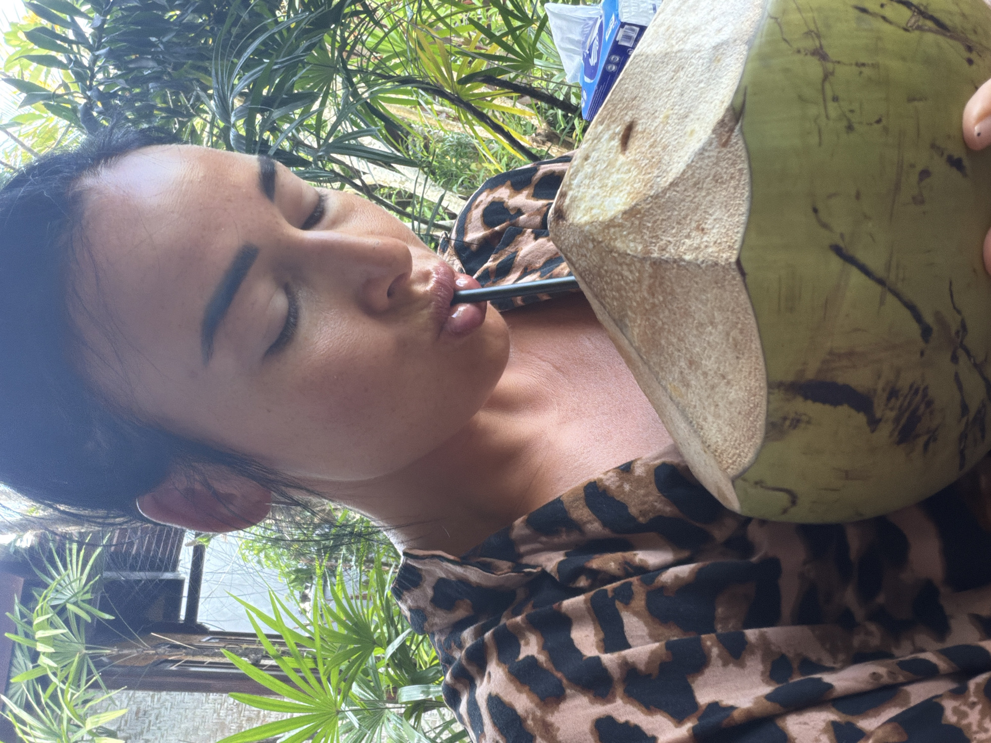 A medium, high-angle shot depicts a person with dark hair, tanned skin, and closed eyes drinking from a green coconut with a straw. The individual is wearing a brown and black animal print shirt. The backdrop features a green, leafy environment, along with a partially visible building with a woven exterior, and a box of tissues. The location is Kutuh, Indonesia.