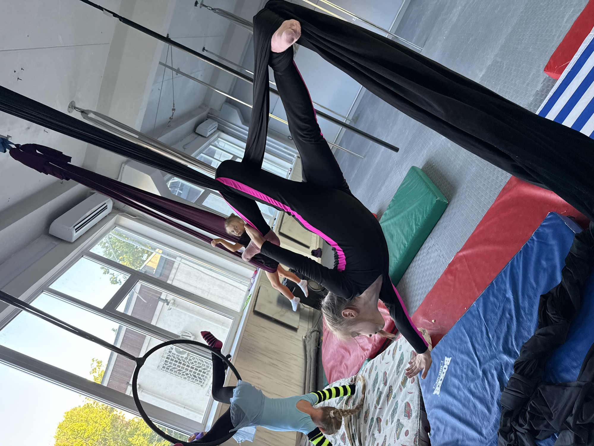 The image shows a group of young girls practicing aerial acrobatics in a studio. Three girls are visible, all engaged in various poses with aerial silks and a hoop. One girl in the foreground, wearing a black leotard with pink trim and pink socks, is suspended upside down, with her legs wrapped in black aerial silks. Her face is turned away from the camera. To her left and slightly behind her, another girl in a light blue leotard and black and yellow striped socks is positioned on an aerial hoop. She is also in an upside down pose, with her legs extended. Further back and to the left, a third girl, wearing a dark outfit and pink socks, is suspended in the air with her legs bent. The studio has a high ceiling with visible metal beams and lighting fixtures. Large windows are visible on the left, offering a view of trees and buildings outside, suggesting it is daytime with bright sunlight. Several colorful mats, including red, blue, and green, are spread on the floor, likely for safety. The atmosphere appears to be one of focused training and athletic activity. The visible text on one of the blue mats reads "BOXER". The location context is Dnipro, Ukraine.