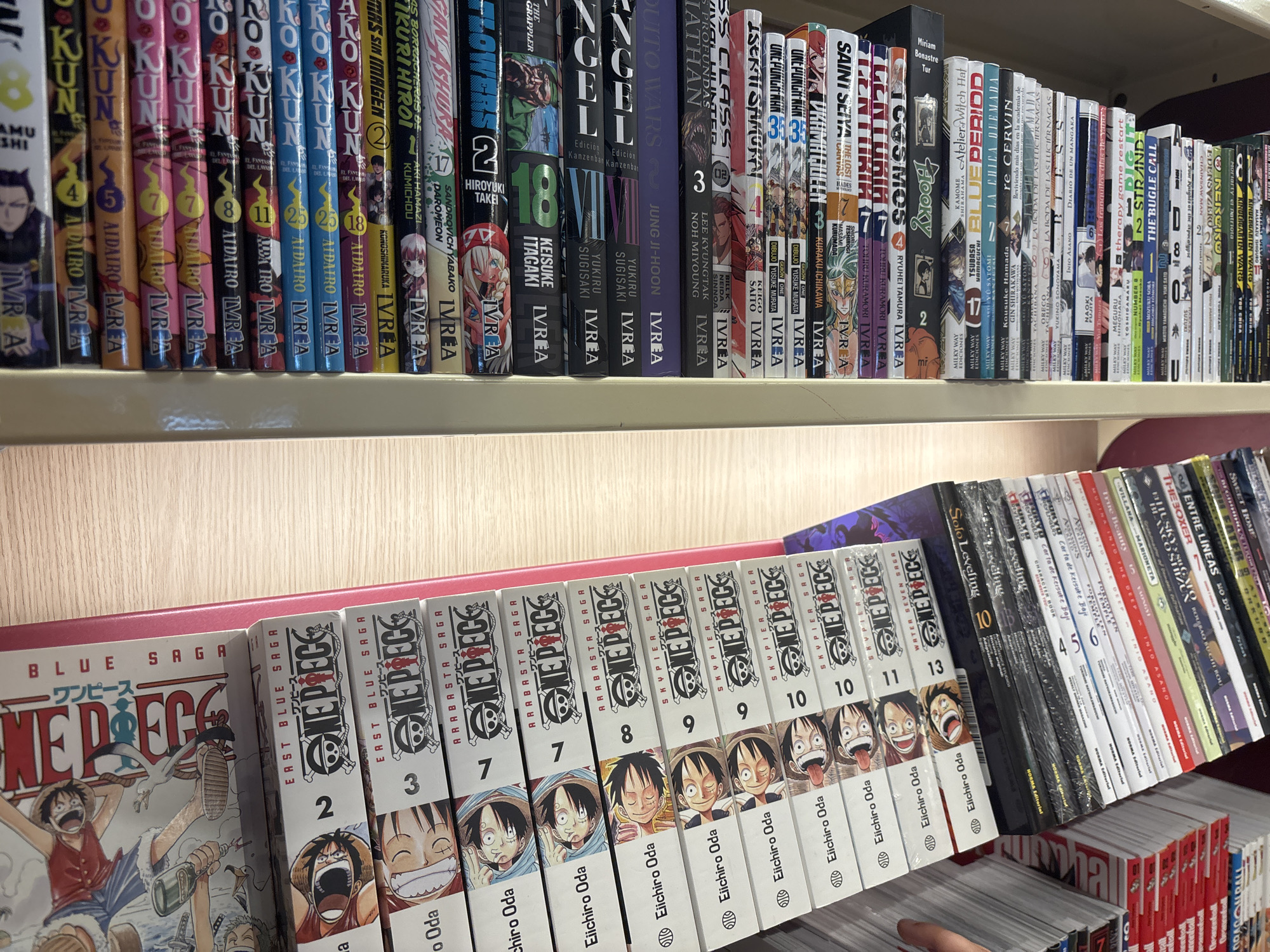 The image displays a shelf filled with manga books. On the top shelf, a collection of manga volumes, predominantly with Japanese titles and some with English and Spanish text, are arranged. These volumes feature a variety of cover art and designs. Some titles visible include "DAIRO," "ANGEL," and "JUNG JI-HOON." On the lower shelf, a prominent set of "ONE PIECE" manga volumes are stacked, identifiable by their distinct cover art featuring characters from the series and the "ONE PIECE" logo. These volumes are labeled with numbers and "Eiichiro Oda" as the author. Interspersed with the "ONE PIECE" manga are other manga titles, some with Spanish text, such as "Solo Leveling," and various other books. The lighting suggests an indoor setting, likely a retail environment or a personal collection. The overall scene is a detailed view of a bookshelf packed with a diverse selection of manga. There are no people or overt actions visible in the image. The location is a shelf in a store or home.