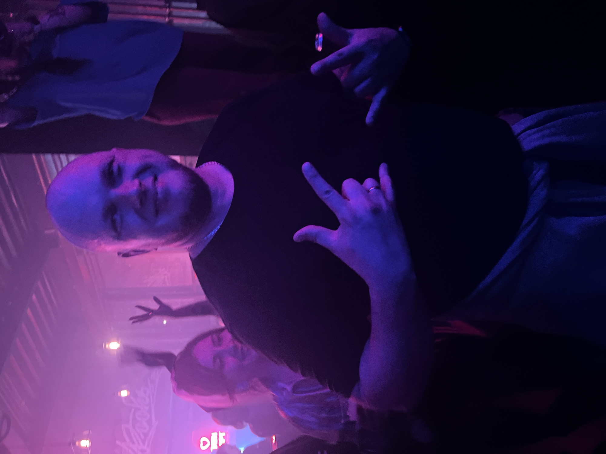 A bald man in a black shirt is in a dimly lit environment, illuminated with purple and pink lights. He is holding up his hands in a "rock on" gesture. There are other people visible in the background. A neon sign is partially visible in the lower center of the frame. The location is the city of Unknown, Unknown.