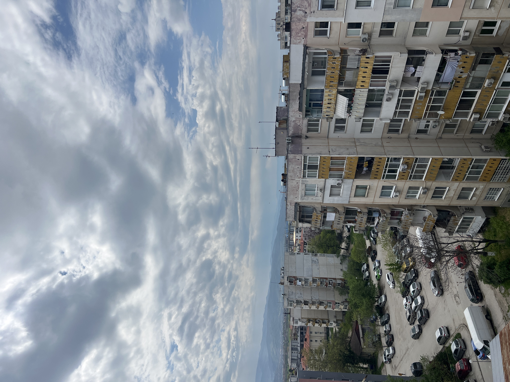 The image provides a high-angle view of a residential area in Plovdiv, Bulgaria, captured on a cloudy day. The scene is dominated by a mix of urban architecture and an expansive sky.
The upper portion of the frame is filled with a dynamic cloudscape, featuring a blend of white, wispy clouds and denser, grey formations, with several patches of clear blue sky visible through the breaks.
On the right side of the image, a series of multi-story apartment buildings stand prominently. These concrete structures show signs of age and wear, with visible peeling paint, discolored facades, and various modifications such as enclosed balconies, numerous air conditioning units, and satellite dishes. The windows are varied, reflecting the diverse living spaces within.
Below and between these buildings, a dense parking area is visible, filled with a multitude of cars of various makes, models, and colors, including sedans, hatchbacks, and a white van. Several trees, some bare-branched and others showing nascent green foliage, are interspersed among the parked vehicles and along the street, providing natural elements within the urban setting.
In the distant background, a range of hazy mountains or hills stretches across the horizon, adding depth to the landscape. The lighting suggests it is daytime, possibly morning or afternoon. The overall impression is that of a bustling, established urban environment. No discernible text is visible on buildings, signs, or vehicles.
