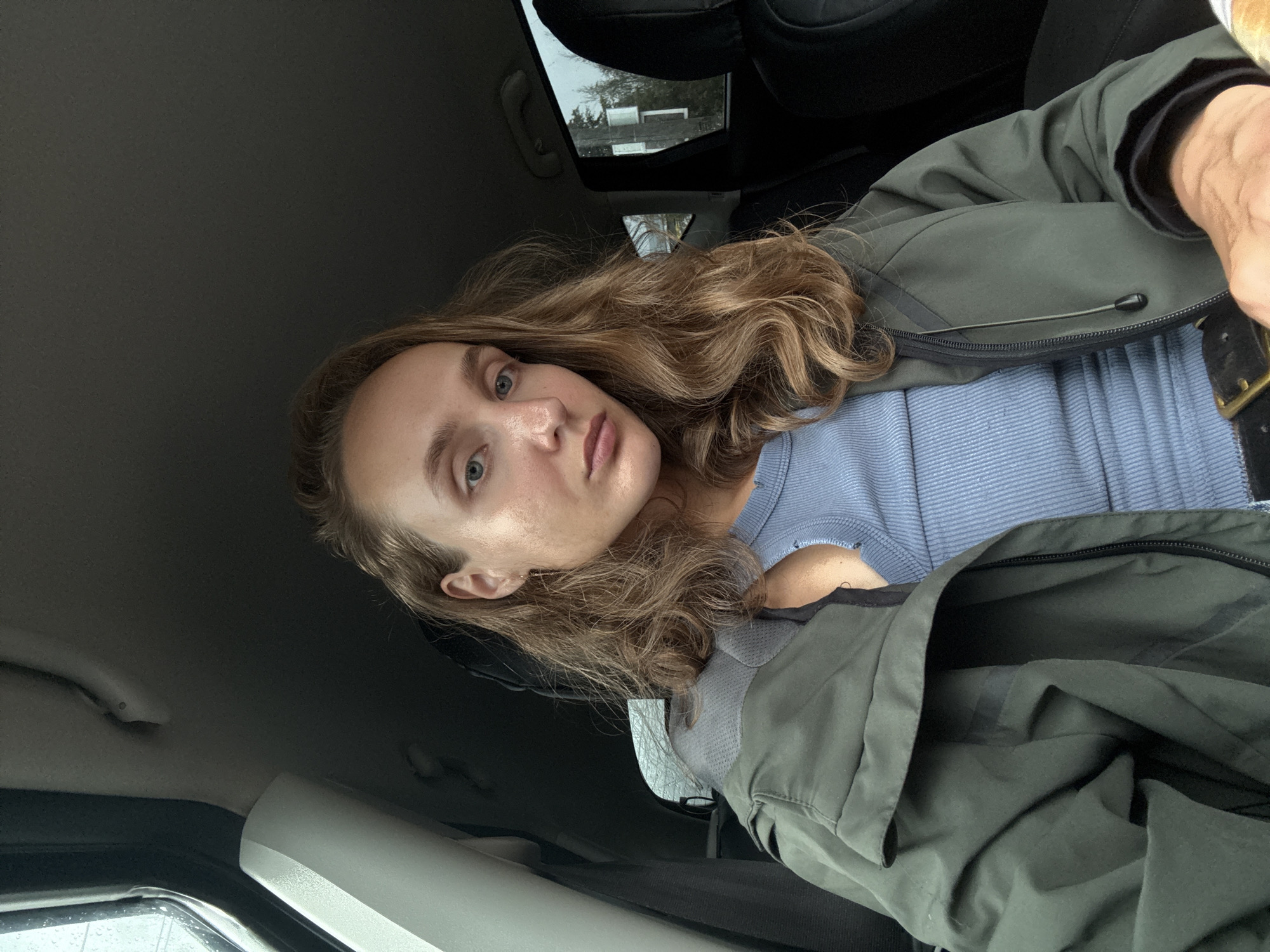 A young woman with long, wavy blonde hair is sitting in the passenger seat of a car, looking directly at the camera. She is wearing a light blue ribbed tank top with a dark green jacket partially unzipped over it. The interior of the car is visible, including the car's ceiling, a window on the left with raindrops on it, and the back of a dark car seat on the right. Outside the window on the right, trees and a pole are visible against an overcast sky, suggesting a cloudy or rainy day. The background appears to be an outdoor setting, possibly a road or a residential area, which could be located in or near the village of Podshivalovo, Russia, given the context. The woman's expression is neutral.