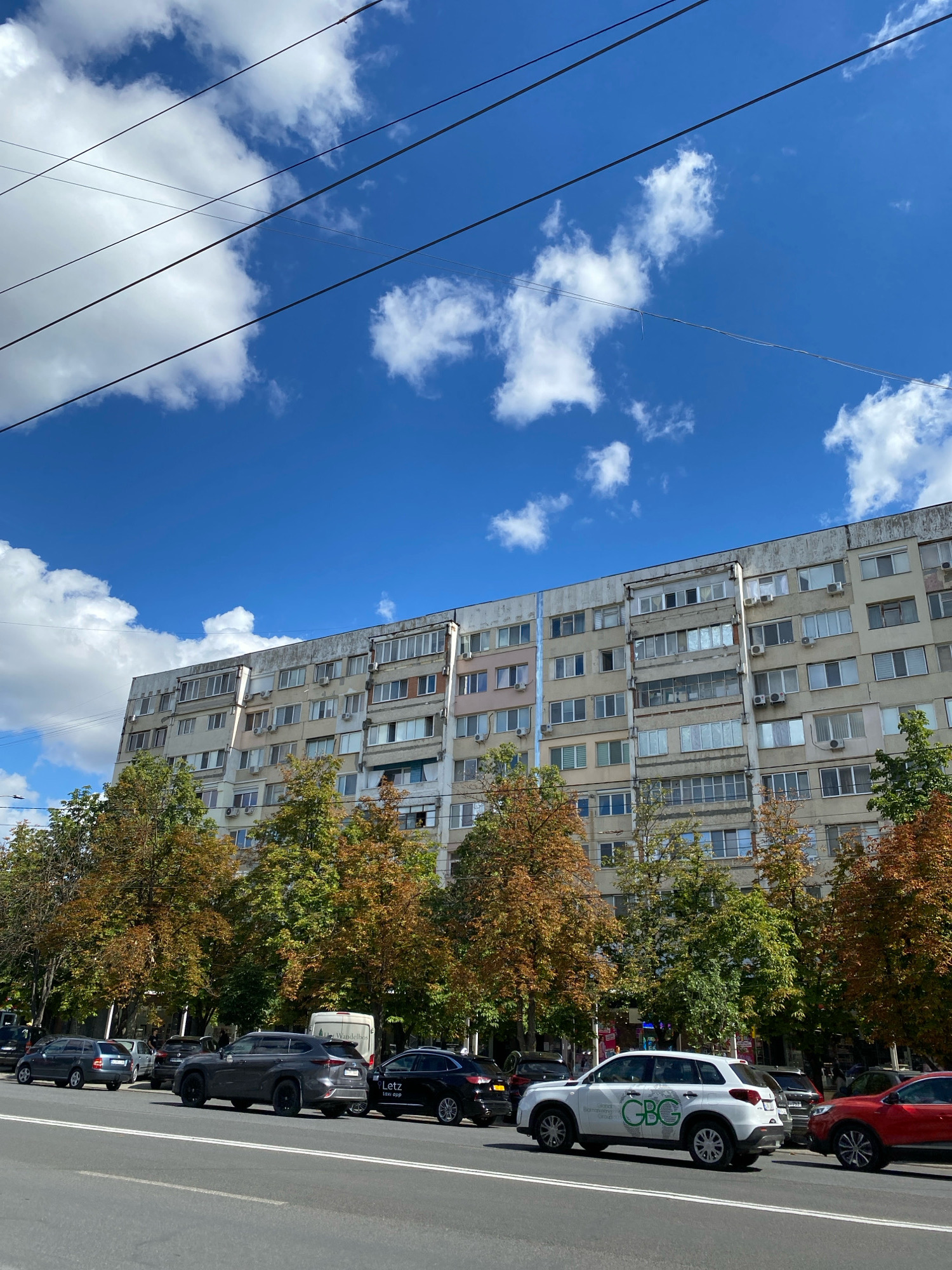 The image depicts a wide street in Chișinău, Moldova, with a large, multi-story apartment building dominating the background. The building is utilitarian in design, with numerous windows and balconies, and shows signs of age. In front of the building, a line of trees displays autumn foliage, with colors ranging from green to yellow and orange. The street is lined with parked cars on the side, and several vehicles are driving in the foreground. The sky is blue with scattered white clouds, and several power lines are visible overhead. The scene appears to be a typical urban environment on a clear day.