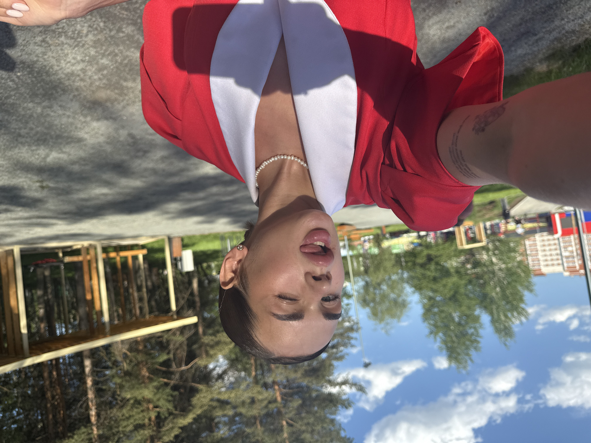 The image shows a selfie of a woman in Perm, Russia. She's wearing a red blazer with a white collar and a pearl necklace. She's winking and sticking out her tongue. Her arm has a visible tattoo. In the background, there are trees, a partially constructed wooden structure, and a building with a Russian flag. The sky is blue with scattered clouds.
