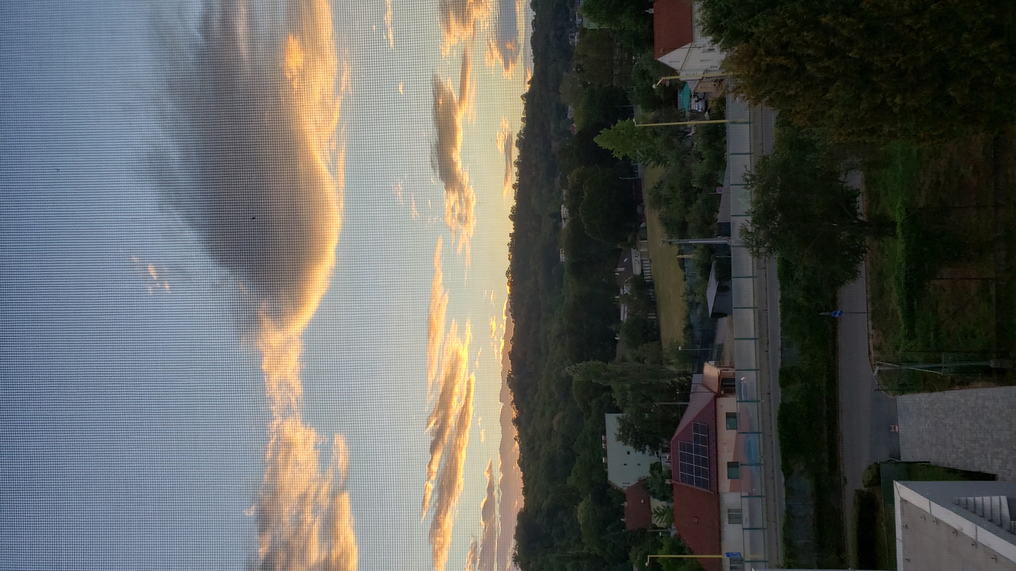 The image depicts a twilight sky with golden-tinged clouds above a tree-covered hillside and a suburban landscape. The view is framed by a window screen. In the foreground, a pathway leads down from a building. Further back, houses with red roofs, one equipped with solar panels, are visible, along with a road and a grassy field. A tall, thin tree stands out in the middle ground. The sky is a blend of pale blue and streaks of orange and yellow, suggesting the sun is setting or has recently set. The overall scene is serene and captures a moment of natural beauty over a residential area.