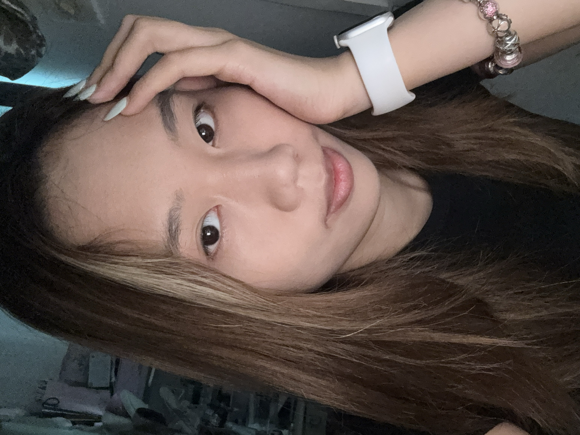 A medium shot captures a young Asian woman from the shoulders up, posed with her right hand touching her forehead and left cheek. She has long, straight brown hair with blonde highlights, framing a face with dark eyes and a slight smile. She is wearing a black t-shirt, a white smartwatch on her left wrist, and a charm bracelet adorned with pink and silver beads. The background is dimly lit, with a teal-colored wall on the left and a dark gray wall on the right. The scene appears to be indoors, possibly in a bedroom or a dimly lit living space in Nan'an District, China.