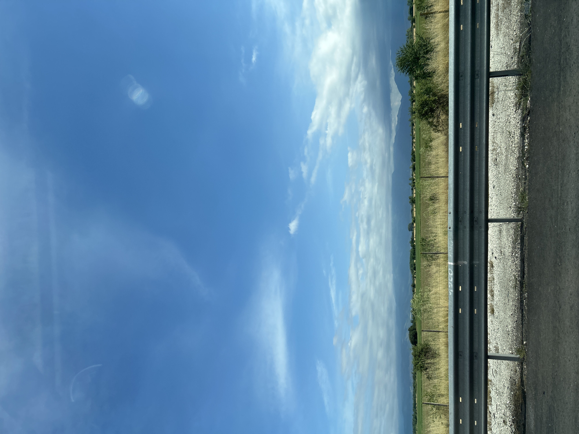 The image captures a roadside scene on a bright, partially cloudy day in Malo Konare, Bulgaria. The primary subject is a multi-tiered metal guardrail, dark grey with reflective horizontal strips, stretching across the middle ground. To the right of the guardrail, a dark asphalt road surface is visible, bordered by a lighter-colored gravel and dirt shoulder. On the guardrail itself, faint, light-colored markings, possibly graffiti, are visible, appearing to spell out "COOL" in the central area.
Beyond the guardrail, to the left, lies an expansive agricultural field with dry, yellowish-green grass and scattered trees and shrubs. In the far distance, under a hazy sky with more substantial cloud cover, the faint silhouettes of mountains or large hills are discernible.
The sky occupies the upper portion of the image, showcasing a vibrant blue with various white cloud formations. These range from thin, wispy cirrus clouds to larger, fluffier cumulus clouds, particularly towards the distant horizon. Several circular and semi-circular lens flare artifacts are present in the upper left quadrant of the sky. The overall impression is one of a calm, open rural landscape viewed from a moving vehicle. No people or specific activities are depicted beyond the implied travel. The time of day appears to be mid-day due to the bright, well-lit conditions.