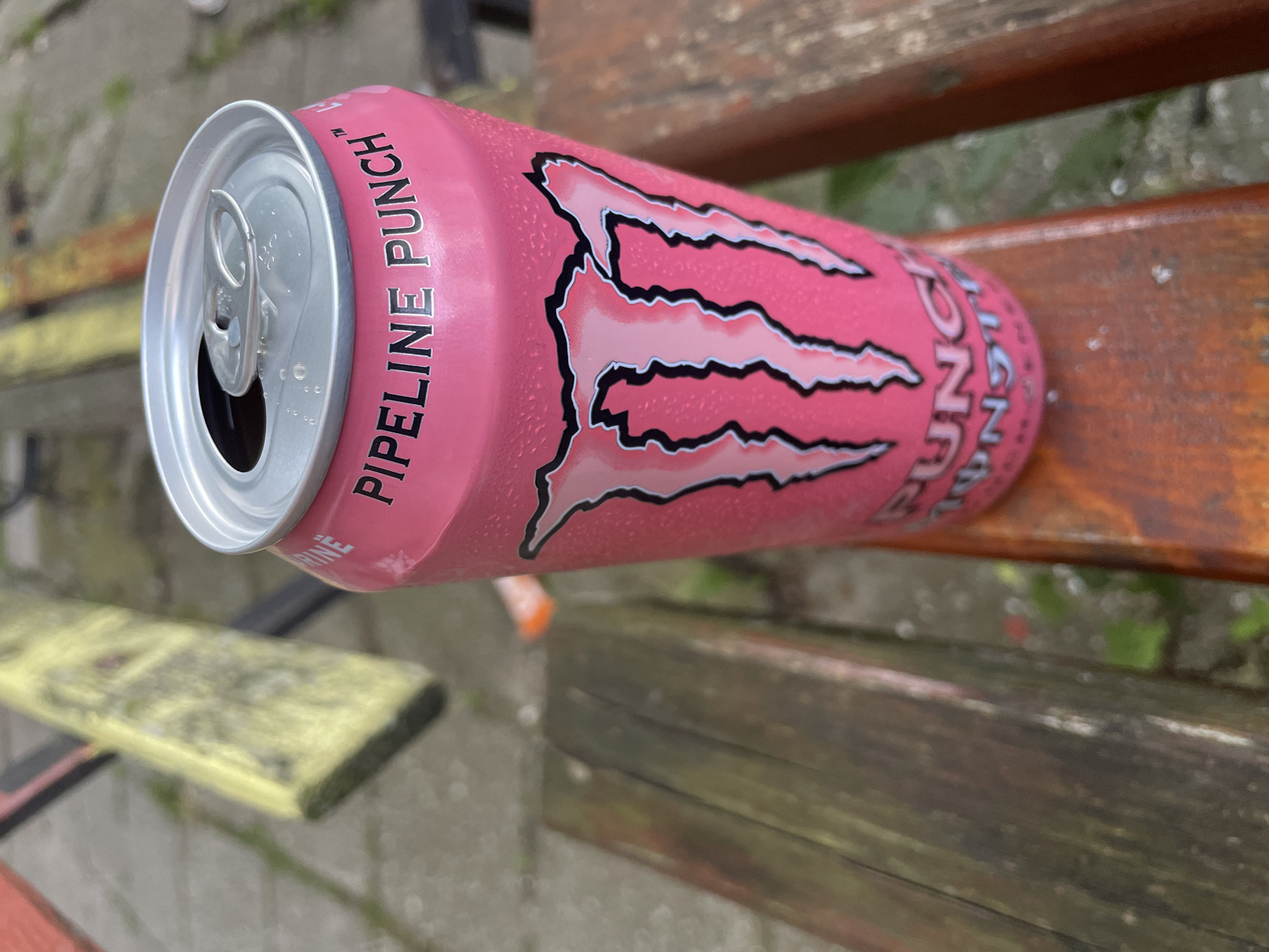 A slightly open can of Monster Pipeline Punch energy drink is lying on a weathered wooden bench. The can is pink with the brand's claw-like logo and the words "PIPELINE PUNCH". The bench has multiple wooden slats, some with yellow paint and others with a worn, brown finish. The background shows a blurred outdoor setting, likely a park or public space. The specific location in Burgas, Bulgaria, is not identifiable from the image.