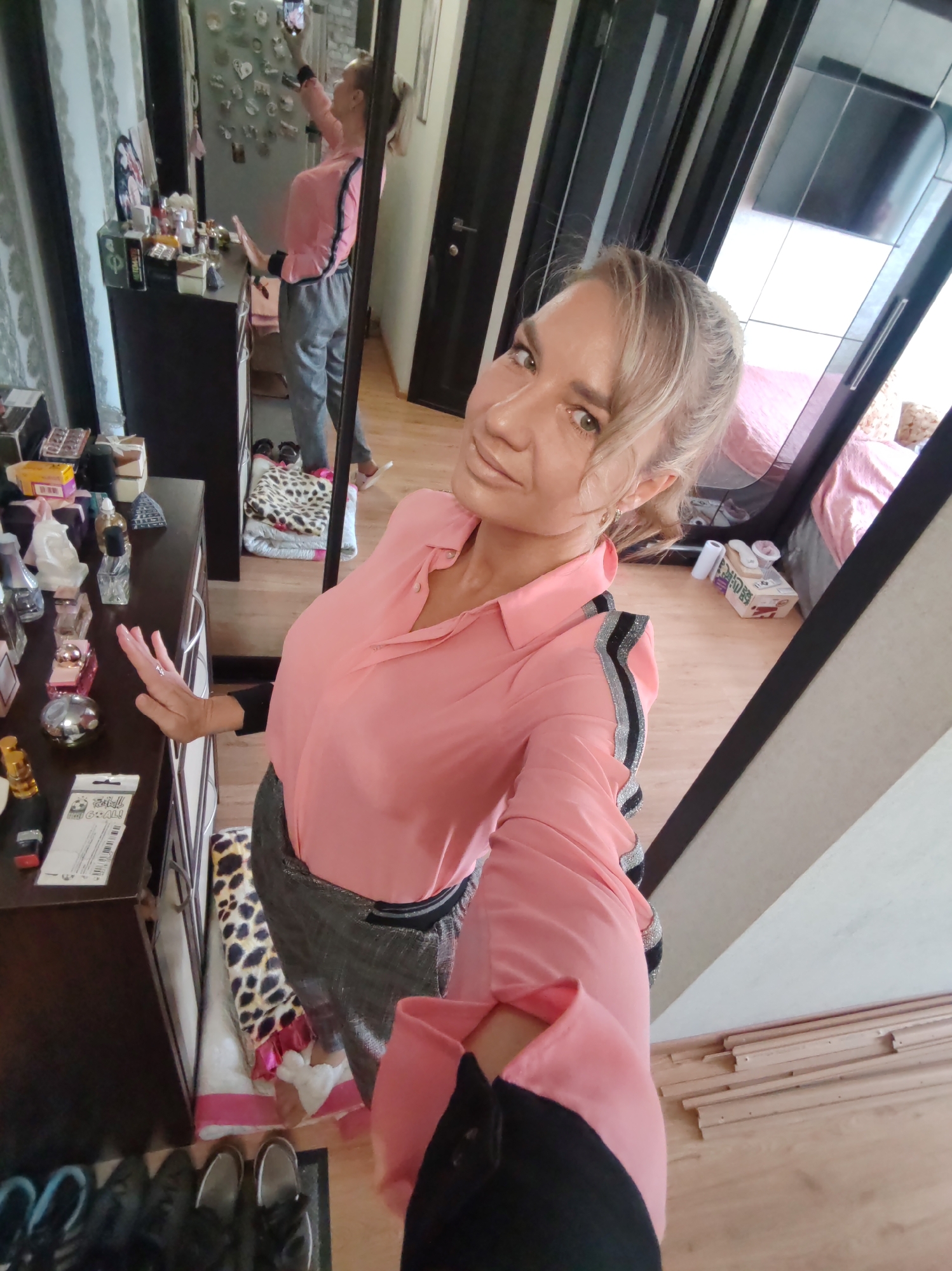This is a selfie taken by a woman in her home. She is standing in front of a large mirror, which reflects her and a portion of the room. The woman has blonde hair pulled back in a ponytail and is wearing a pink collared shirt with striped detailing on the sleeves and a grey skirt. She is holding her phone in her right hand, taking the picture, and her left hand is raised, resting on a dresser.
The dresser is dark brown and cluttered with various items, including perfume bottles, small boxes, and what appears to be a lottery ticket with Cyrillic text. To the left of the dresser, a pile of boxes and bags is visible. In the reflection of the mirror, the woman is seen wearing grey sweatpants and white slides. Behind her, a bed with pink bedding is partially visible, along with a closet with mirrored doors. The room appears to be a bedroom or dressing area, with wooden flooring and light-colored walls. The scene suggests a casual moment, possibly getting ready or taking a quick photo.