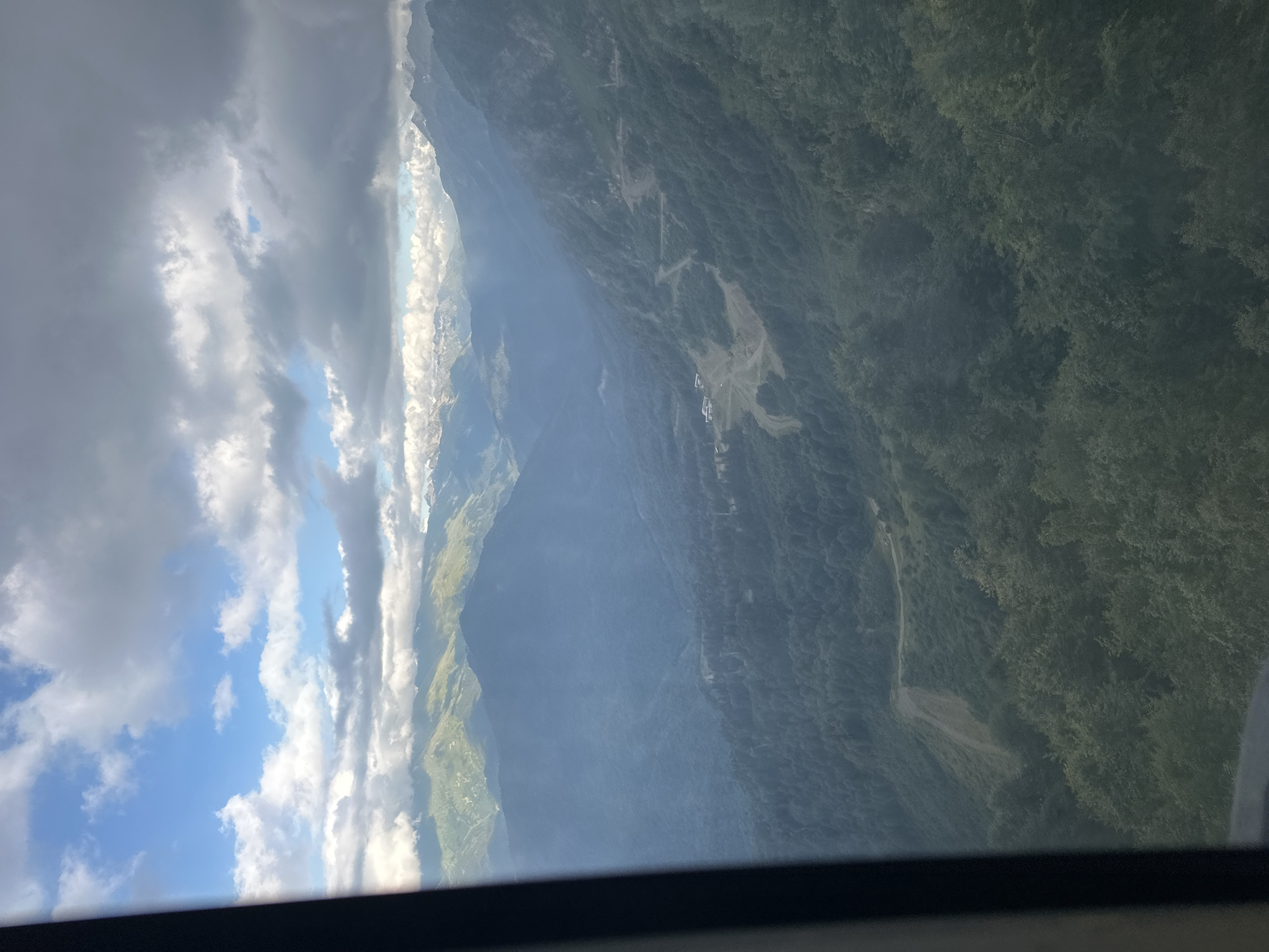 This is an aerial, wide-angle photograph captured from a cable car, looking out over a mountainous landscape in Estosadok, Russia. The scene is dominated by layers of rugged, tree-covered mountains extending into the distance. In the foreground, the dark, blurred edge of the cable car's window frame partially obstructs the view on the left side.
The immediate midground displays a dense, dark green forest covering the slopes. A winding, dirt road is visible snaking its way up one of the mountainsides, and a small cluster of buildings, possibly a resort or service area, is situated in a clearing. The lighting suggests a partly cloudy day, with shafts of sunlight breaking through the overcast sky and illuminating patches of the distant peaks.
Further into the background, the mountains become progressively hazier and bluer due to atmospheric perspective, with some peaks appearing snow-capped. The sky is a mix of bright blue patches and dramatic, dark grey clouds. The overall impression is one of vast, natural beauty and rugged terrain. No people or animals are visible in the frame.