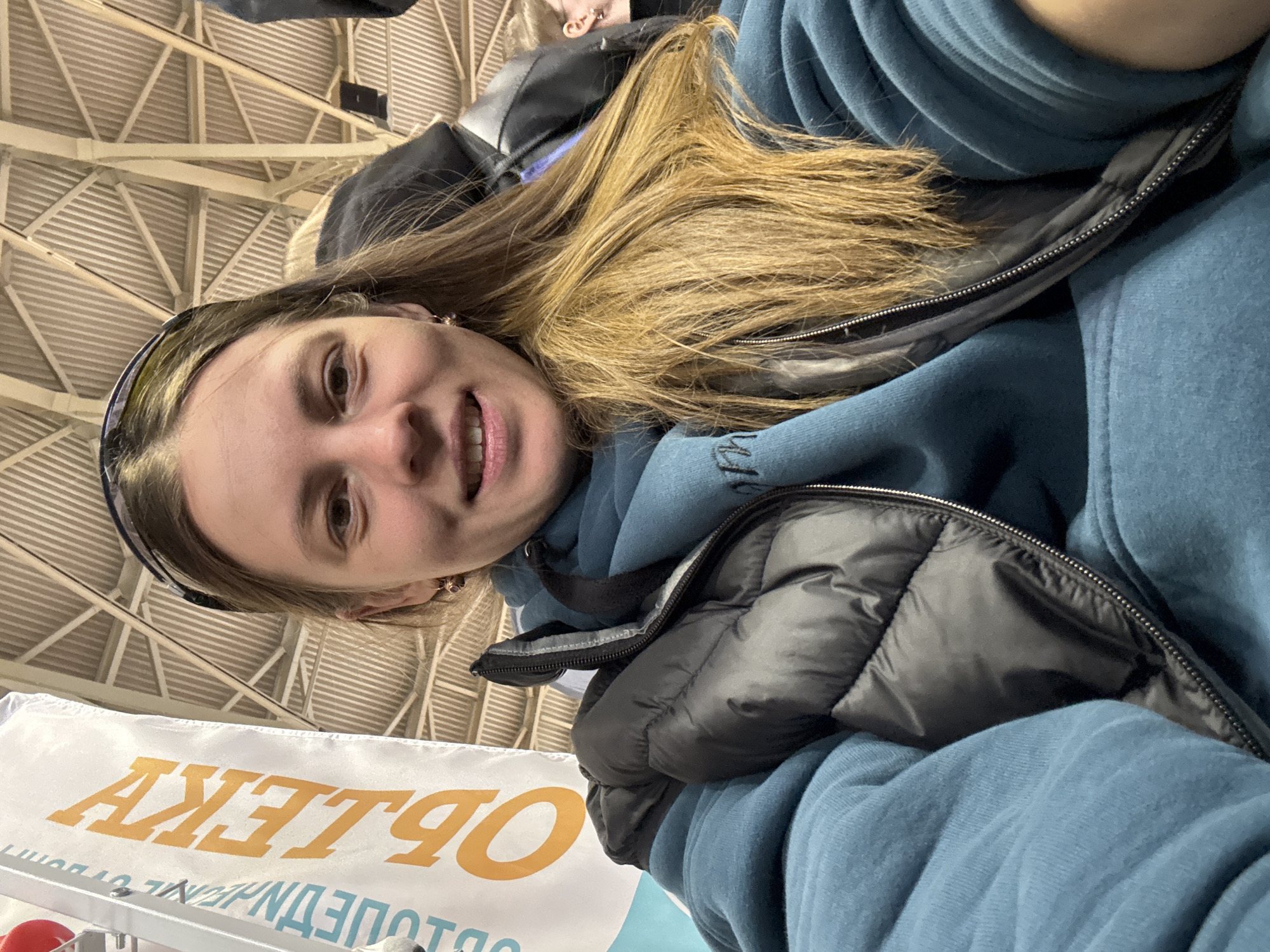 A woman with long, light brown hair, wearing a blue hooded sweatshirt and a black puffer vest, is looking at the camera and smiling. She has a black headband on. Behind her is the ceiling of a large indoor space with a white and metal support structure. In the lower left corner, there is a white banner with orange lettering that reads "ОБЪЕКТЫ". This image was taken in the city of Песчаные Ковали, Russia.