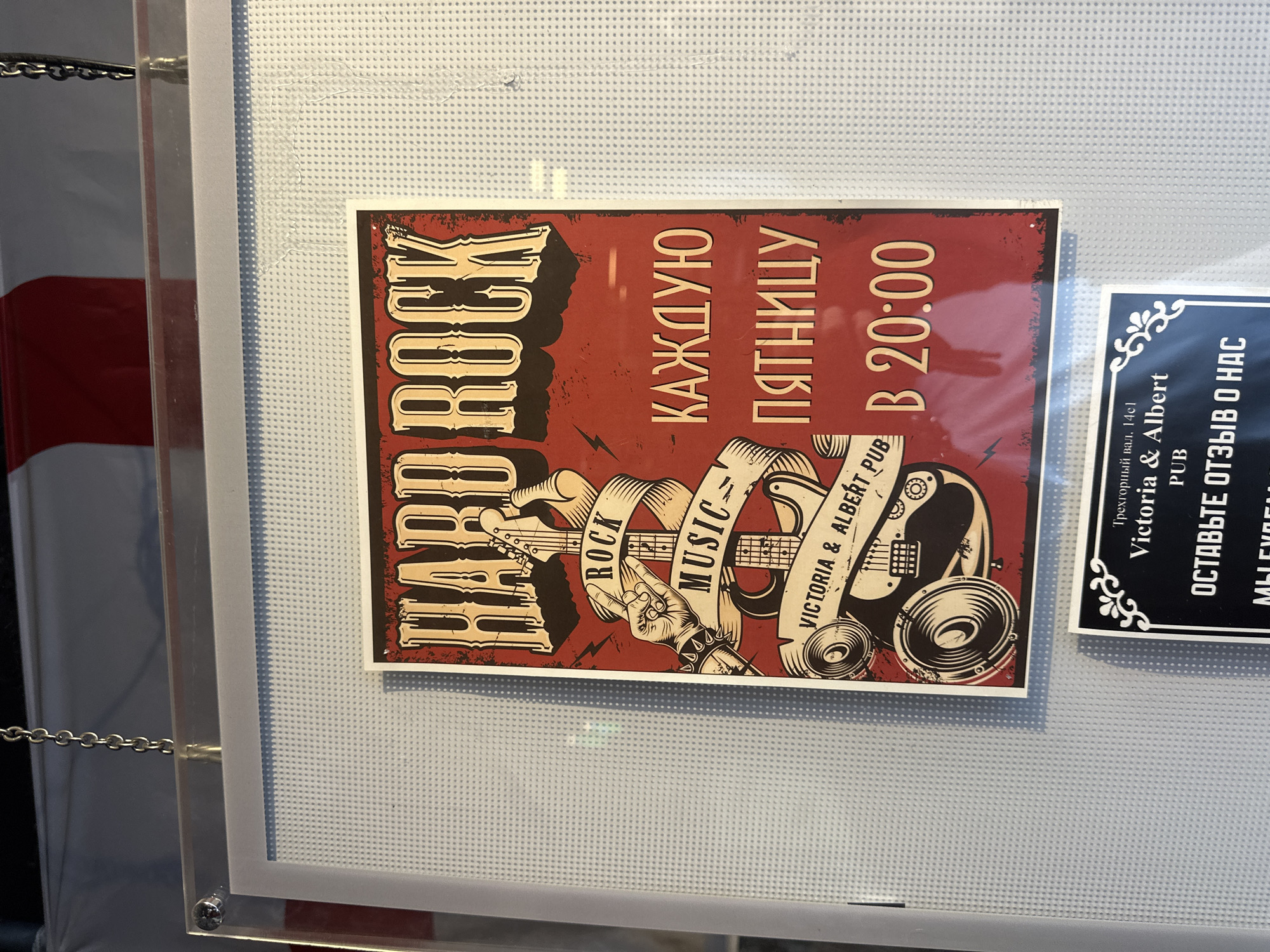 A vertically oriented photograph captures a "Hard Rock" promotional poster displayed behind a clear acrylic panel with a white perforated backing. The poster, predominantly red and black with beige text, features "HARD ROCK" in large distressed font at the top. Below, an illustration depicts an electric guitar, a hand forming a "rock on" gesture with a studded wristband, and two banners inscribed with "ROCK MUSIC" and "VICTORIA & ALBERT PUB". Two large speakers are illustrated at the bottom left. Adjacent to the guitar, in red Russian text, "КАЖДУЮ ПЯТНИЦУ" (Every Friday) and "В 20:00" (at 20:00) are printed. The clear acrylic panel exhibits reflections, including a distorted pink-red and white shape. The display is suspended by two metal chains, with one visible at the top left, attached to a metal standoff. Behind the display, a flag with horizontal red and white stripes is partially visible in the upper background. Below the main poster, a smaller black rectangular sign with white text and decorative flourishes is partially visible. This sign explicitly names the establishment as "Victoria & Albert PUB" and provides the Moscow address "Трехгорный вал, 14с1" (Trekhgorny Val, 14s1), with additional Russian text "ОСТАВЬТЕ ОТЗЫВ О НАС" (Leave a review about us) and partially visible "МЫ БУДЕМ". The scene depicts an indoor environment, indicating a pub or establishment located at Trekhgorny Val, 14s1, in Moscow, Russia.