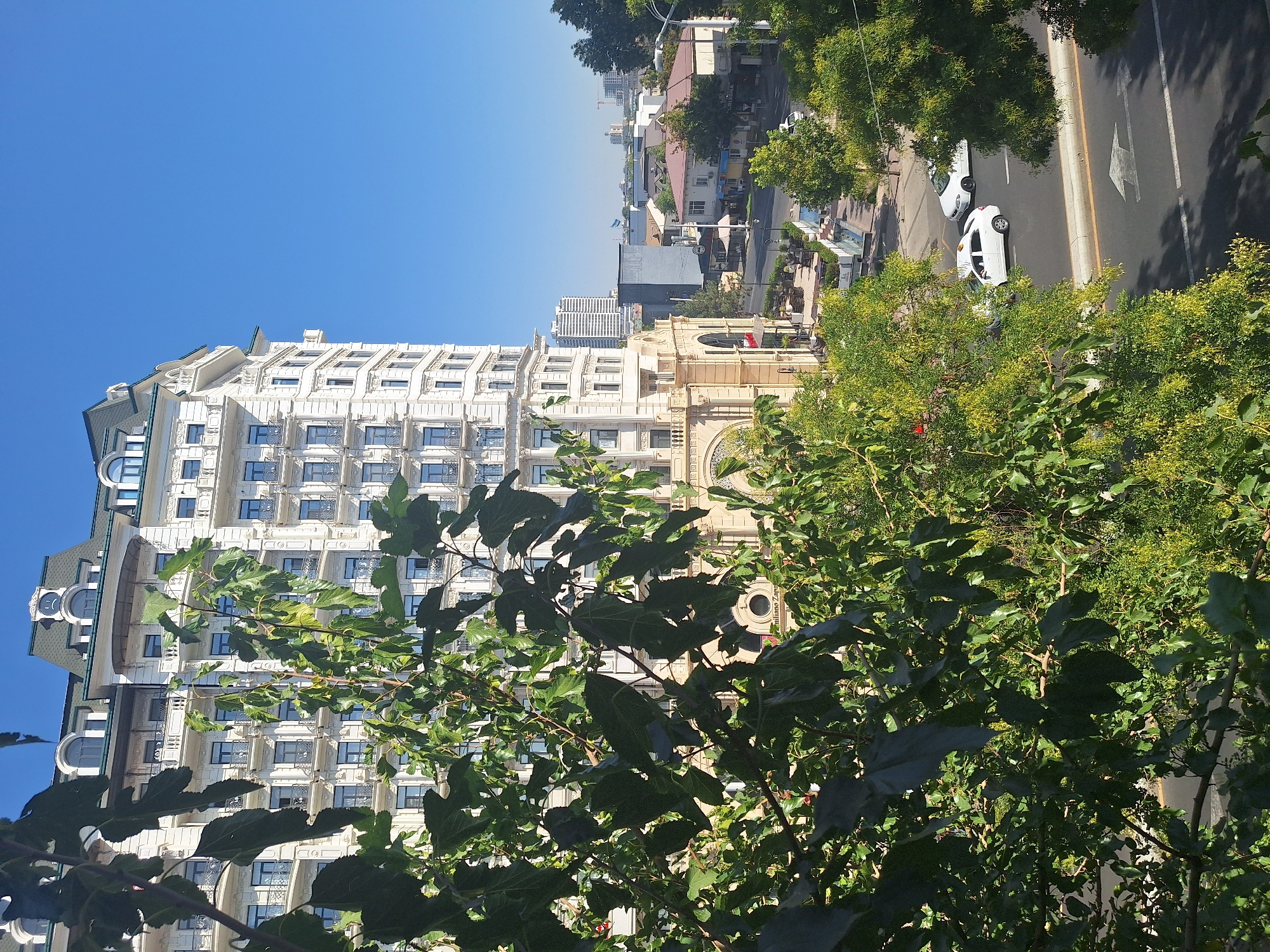 The image captures a sunlit day in Tashkent, Uzbekistan, featuring a grand white building with ornate detailing and numerous windows, partially obscured by lush green foliage in the foreground. The building appears to be a hotel or a prominent commercial establishment, possibly the "Grand Mir Hotel" due to its architectural style, with balconies adorning its facade. In the background, other buildings of varying architectural styles and heights are visible, including a taller, modern high-rise. A street runs along the base of the buildings, with a few white cars driving on it. The sky is a clear, bright blue, suggesting pleasant weather. The overall scene conveys an urban environment with a blend of classical and modern architecture, characteristic of Tashkent.