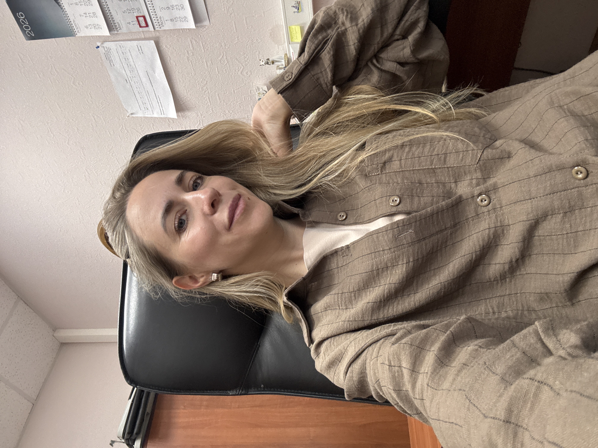 A blonde woman is captured in a selfie-style shot, sitting in a black office chair and looking directly at the camera with a gentle expression. Her long, straight blonde hair cascades over her shoulders and arm. She is wearing a brown long-sleeved shirt with thin darker stripes over a lighter-colored top, and one of her hands is raised towards her head, touching her hair. Small earrings are visible in her ears.
The setting appears to be an office or workplace, indicated by a light-colored, textured wall behind her. On the wall, a calendar from the year "2026" is visible at the top left. Other parts of the calendar show Cyrillic text, likely month names such as "Май" (May) and "Июнь" (June), along with days and numbers. A specific date, possibly "25", is highlighted in red. Below the calendar, a white paper document is pinned to the wall, but its text is unreadable. A dark wooden surface, possibly a desk or table, is visible in the lower part of the image. The ceiling, with white acoustic tiles, can be seen in the upper left corner. The overall lighting suggests an indoor environment.